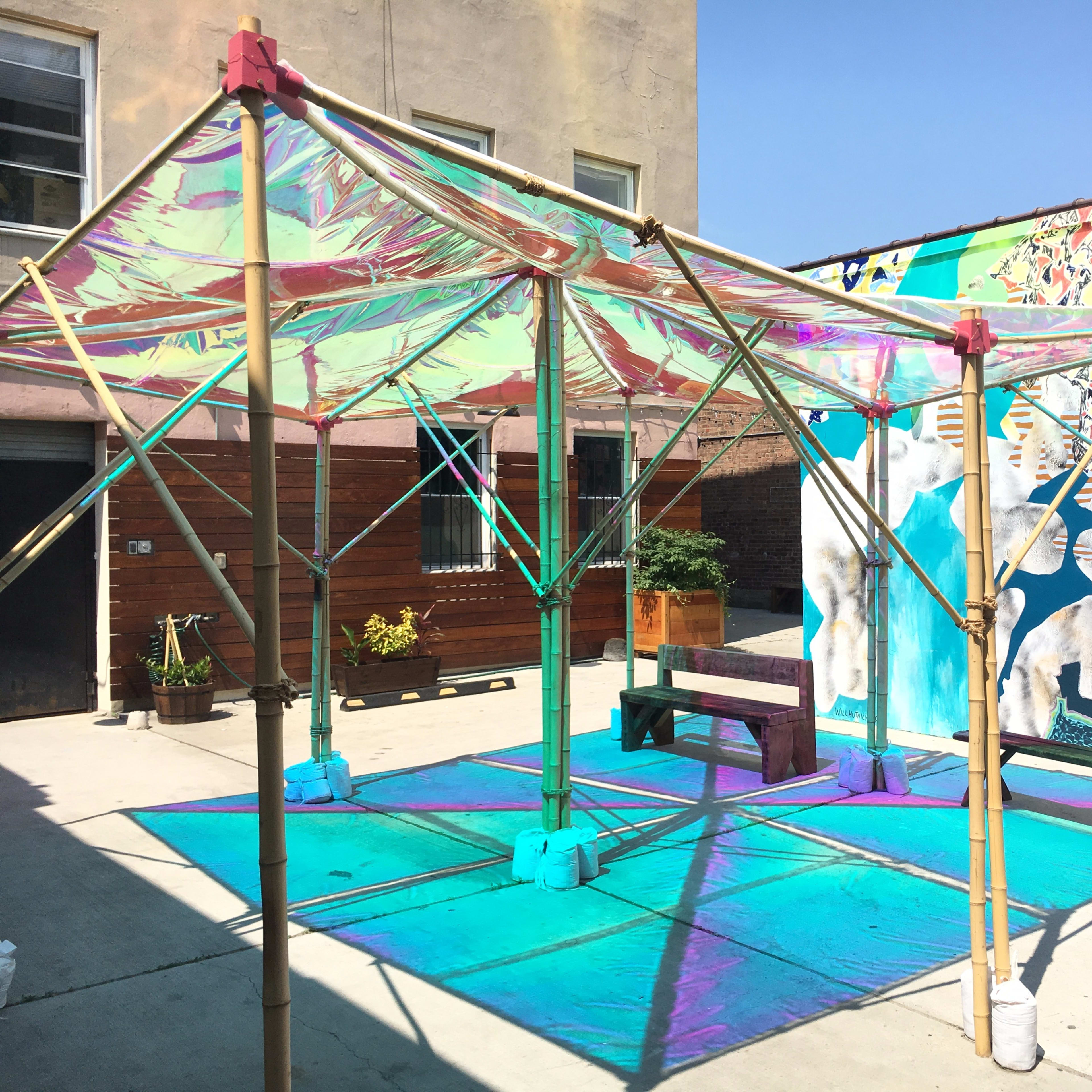 A structure made of bamboo poles and colorful translucent material is set up in an outdoor space beside a mural.