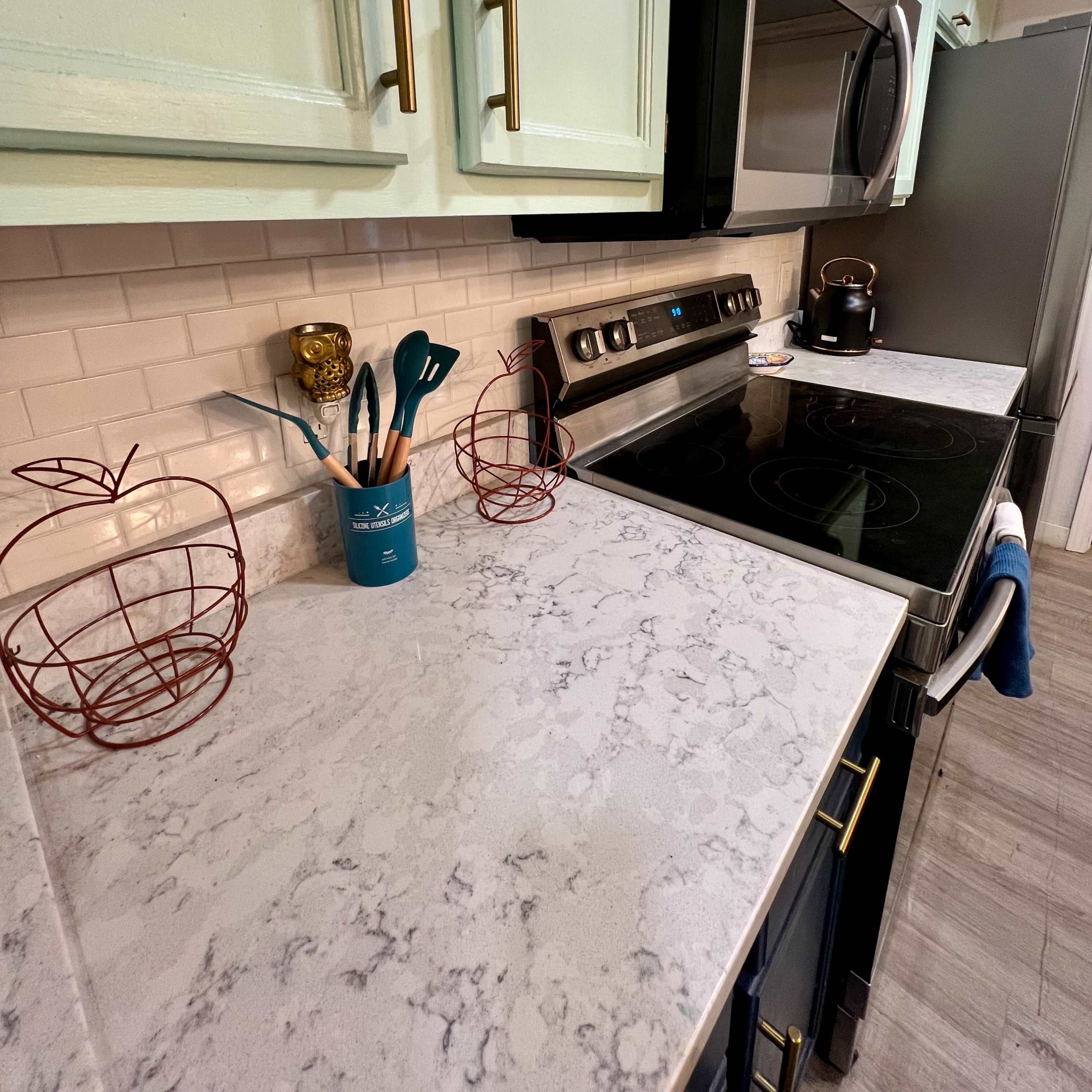 A kitchen countertop with a marble surface, green cabinets, stainless steel appliances, and decorative wire fruit holders.