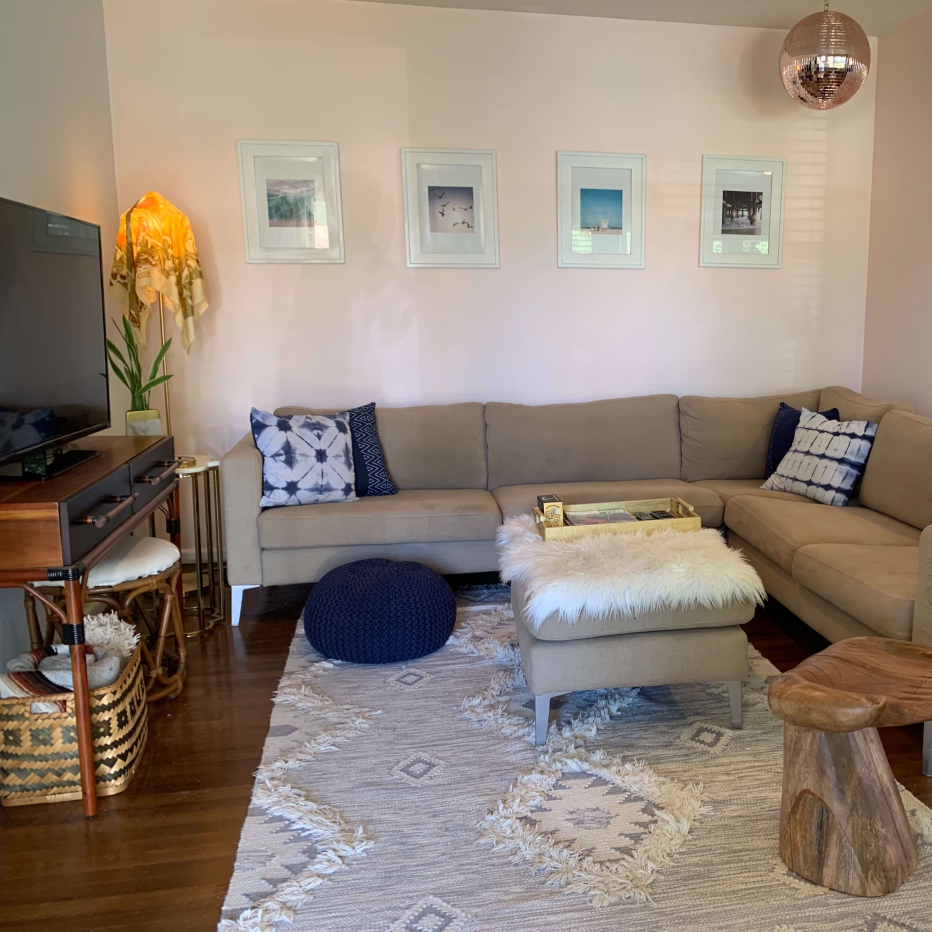 A light-filled living room features a beige sectional sofa, a wooden coffee table, decorative pillows, and a small dog standing on the armrest.
