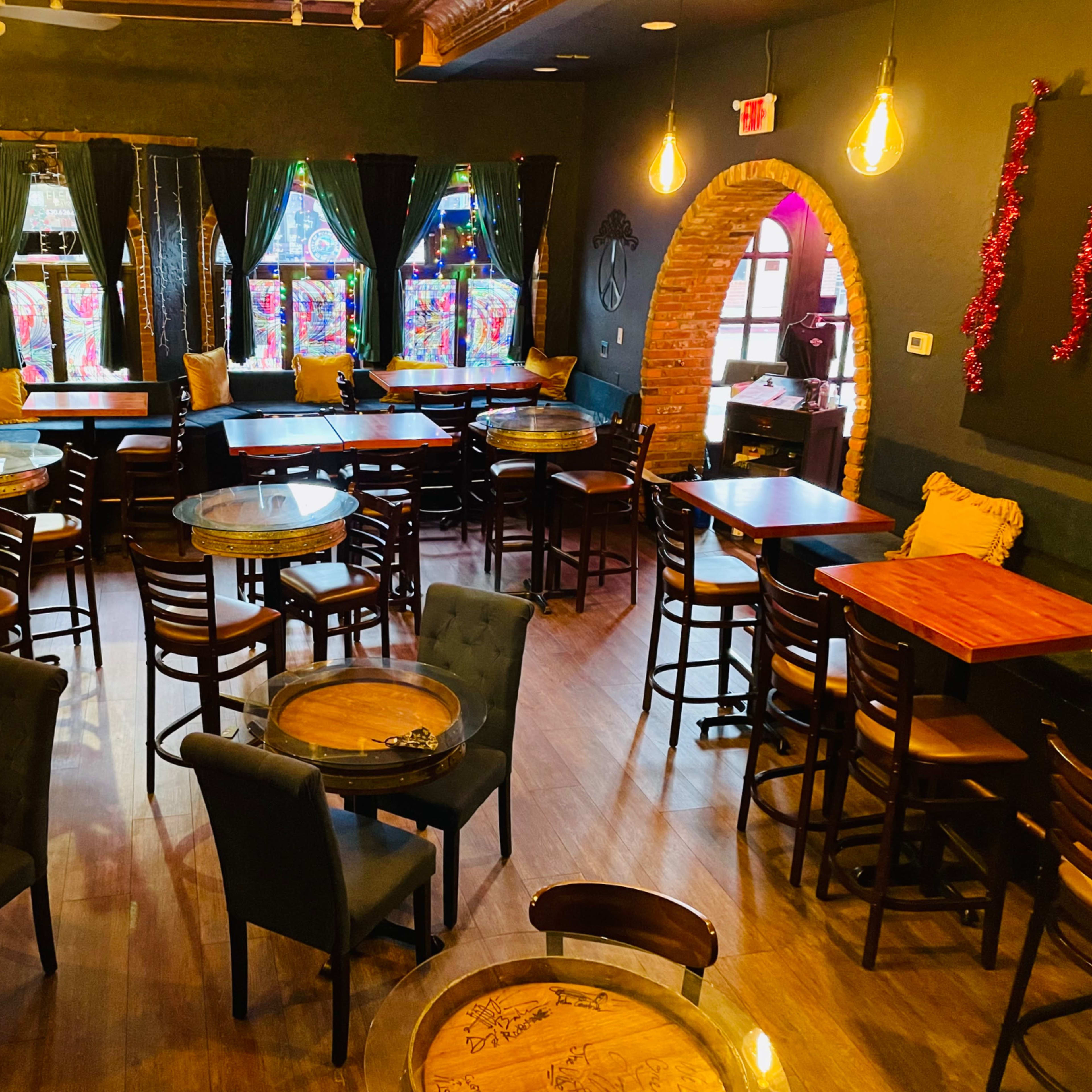 A cozy interior of a café or bar with wooden tables and chairs, large windows featuring colorful stained glass, and dim lighting.
