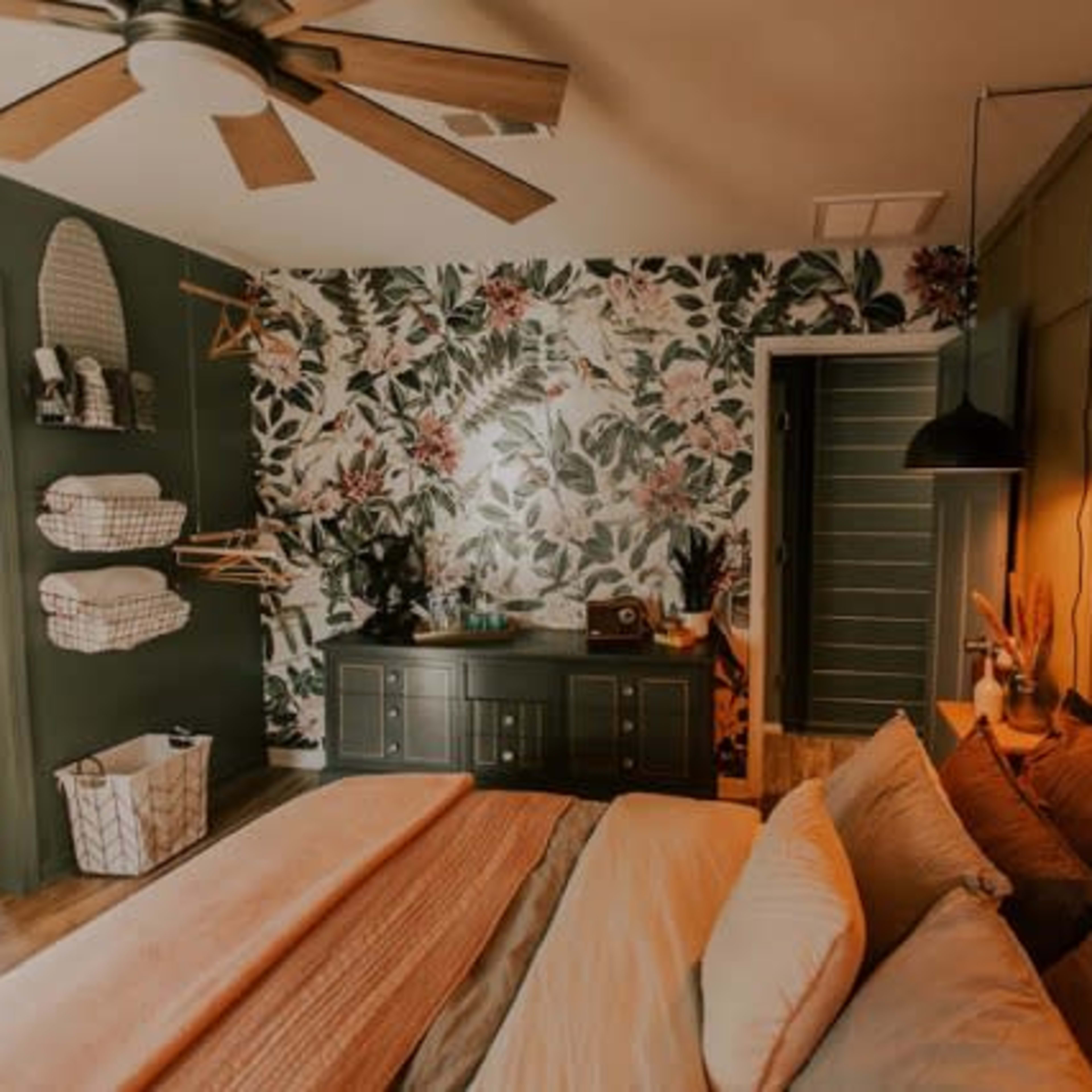A bedroom features a floral accent wall, a wooden dresser, and a bed with layered linens, alongside a door leading to an adjoining bathroom.