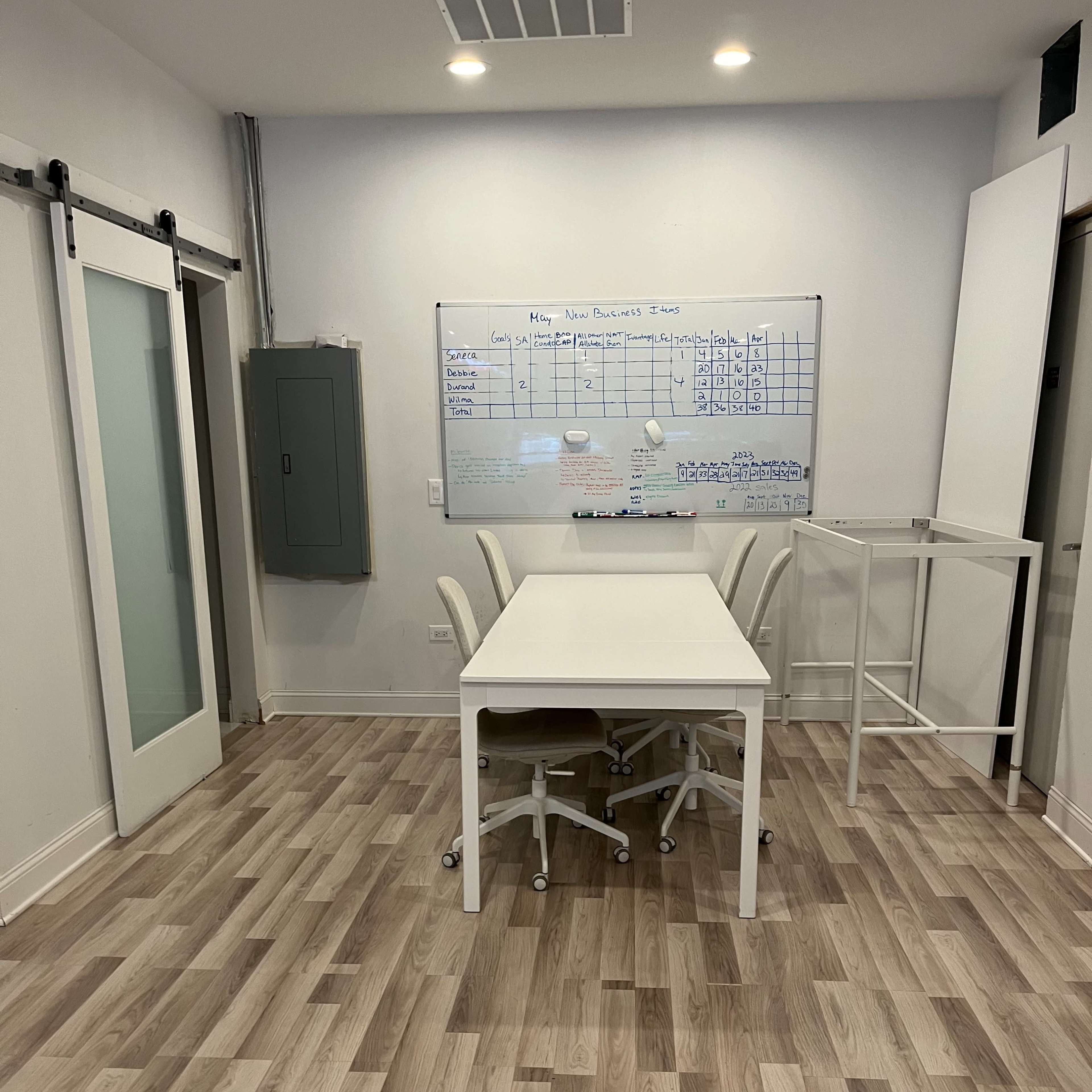 A bright meeting room features a white table surrounded by chairs and a whiteboard with a project schedule.