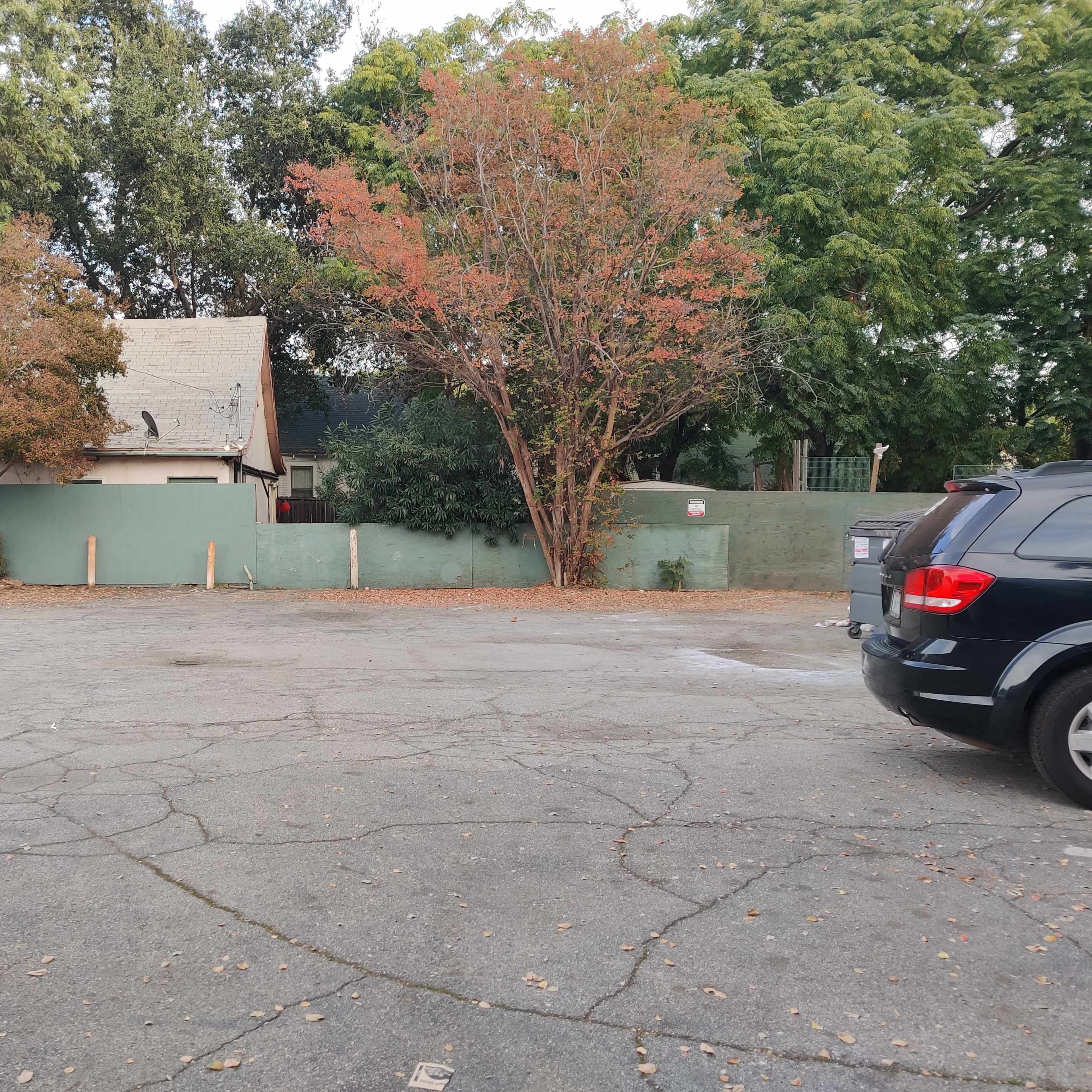 A vacant parking lot with a few trees and a house partially obscured by a green fence in the background.