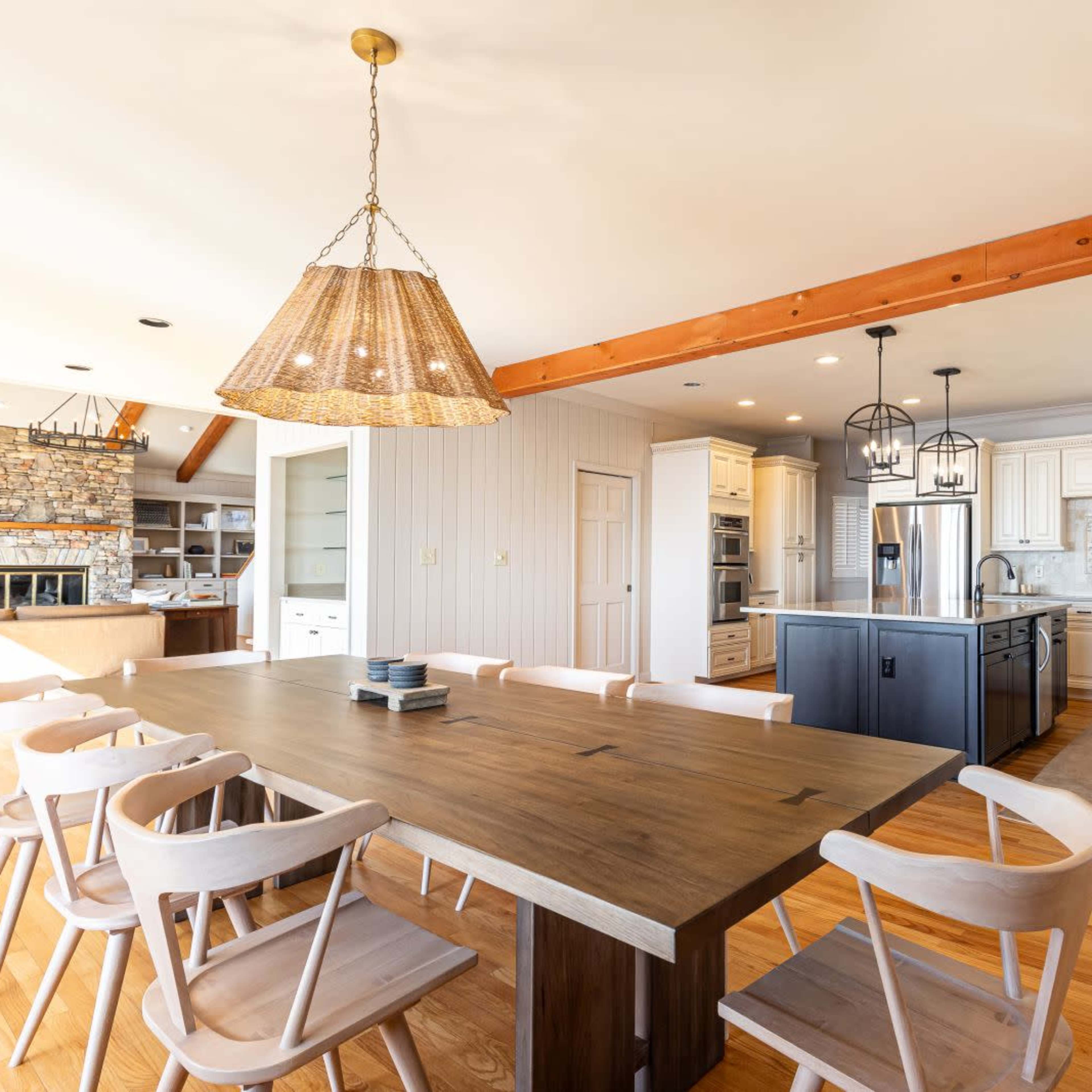 A spacious kitchen features a large wooden dining table surrounded by eight chairs, with a stone fireplace visible in the background.