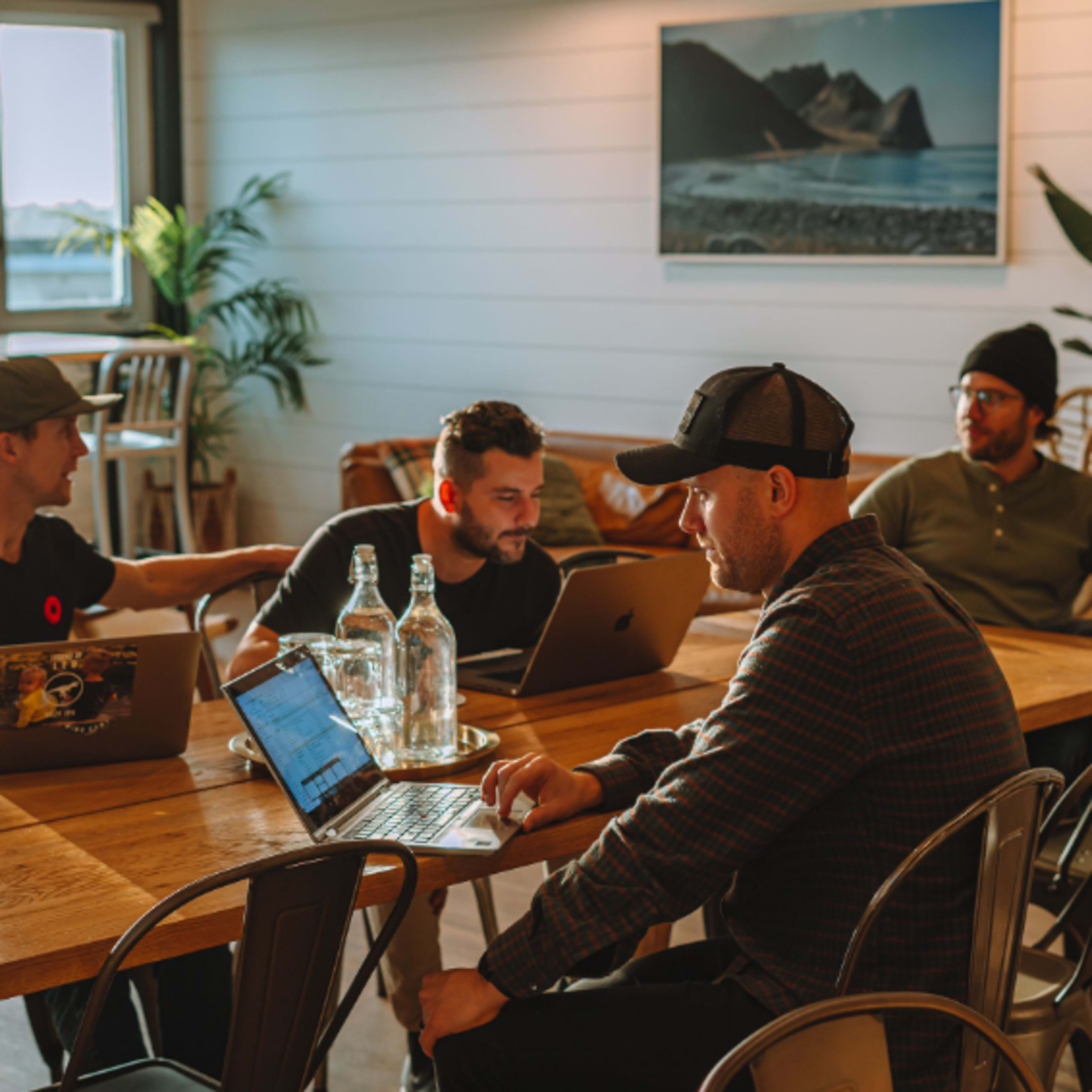 Four individuals work together at a wooden table in a bright, modern workspace with laptops and water bottles.