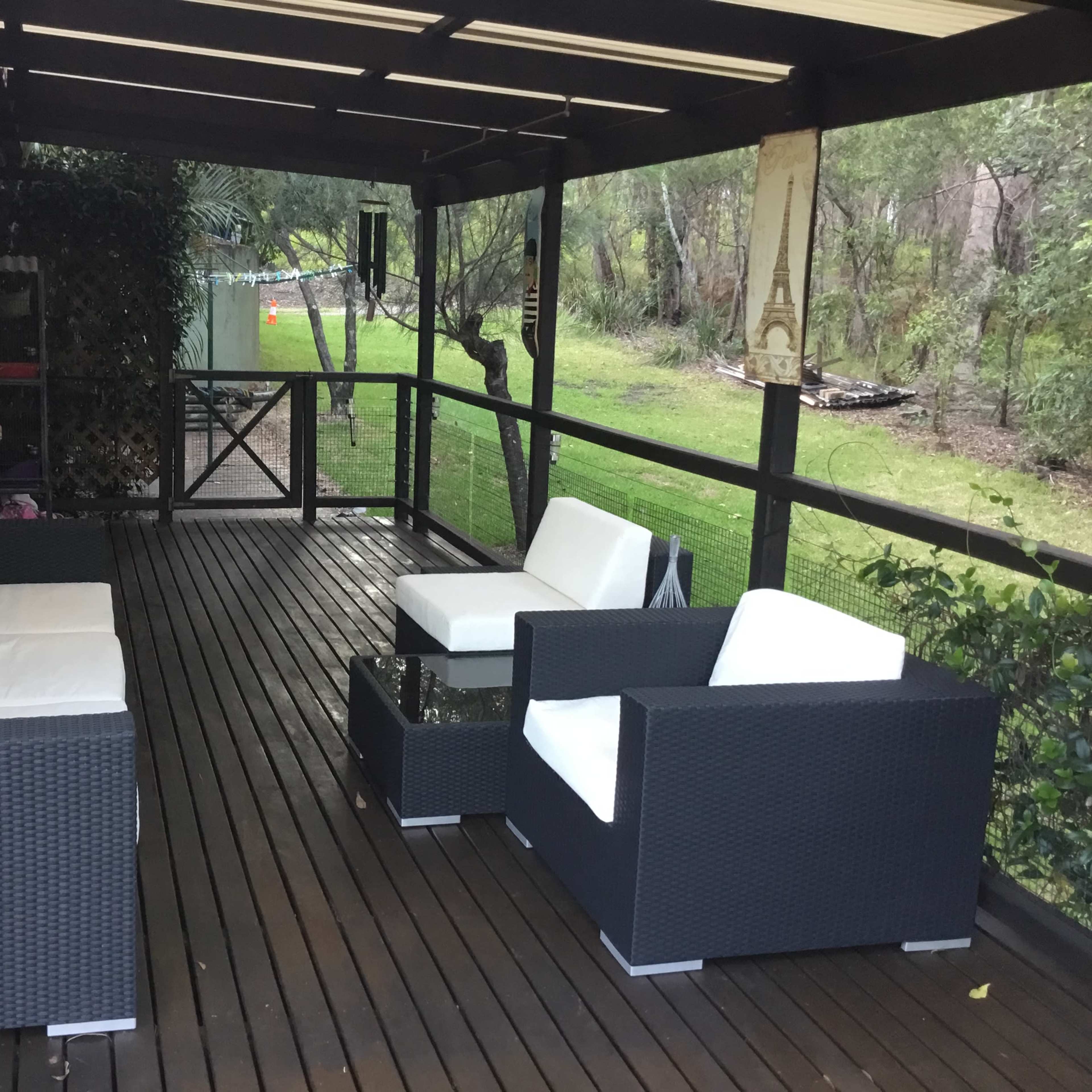 A covered outdoor patio furnished with two white sofas and a glass-top coffee table, surrounded by greenery.