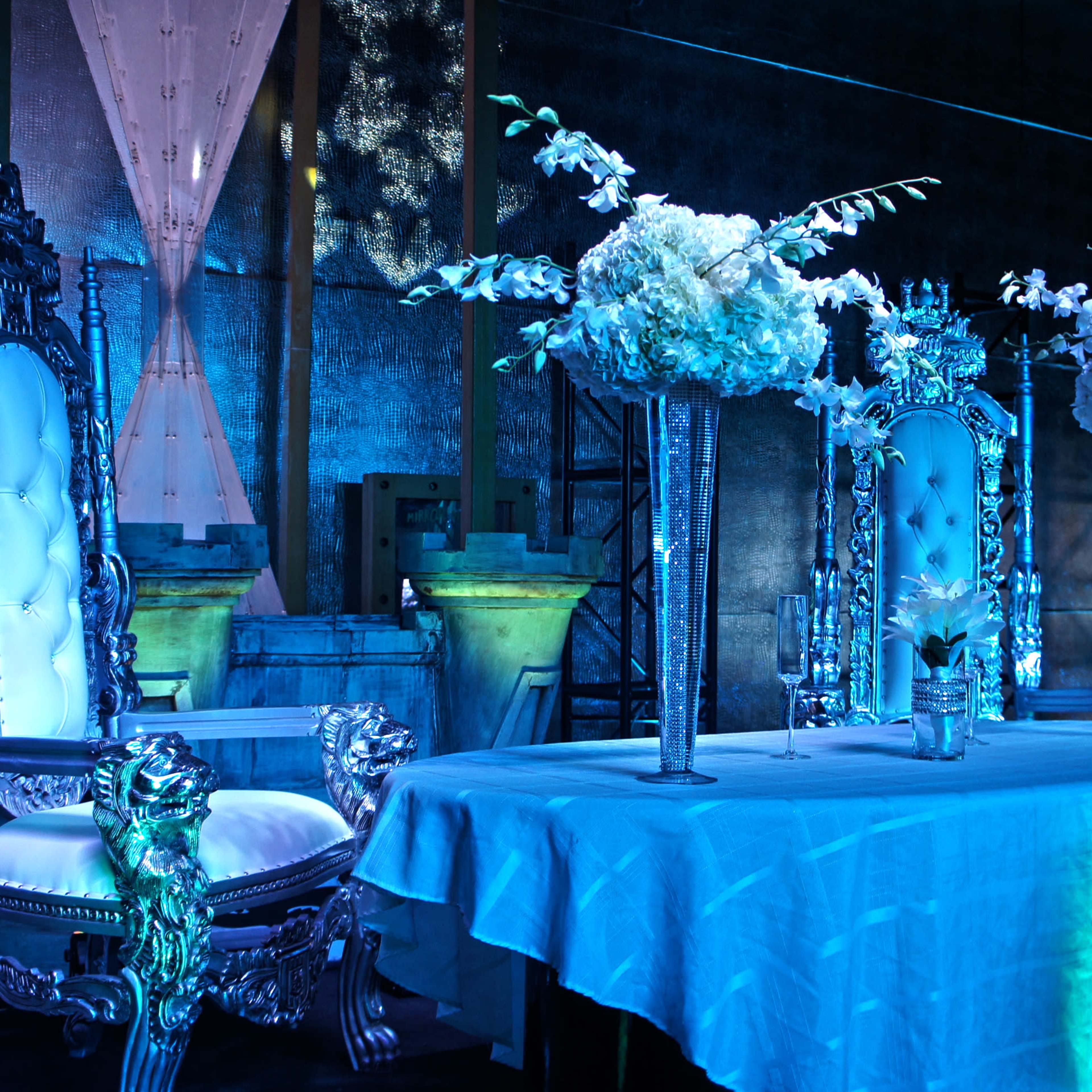 A decorated table with three large floral arrangements is set in front of two ornate chairs in a dimly lit venue.