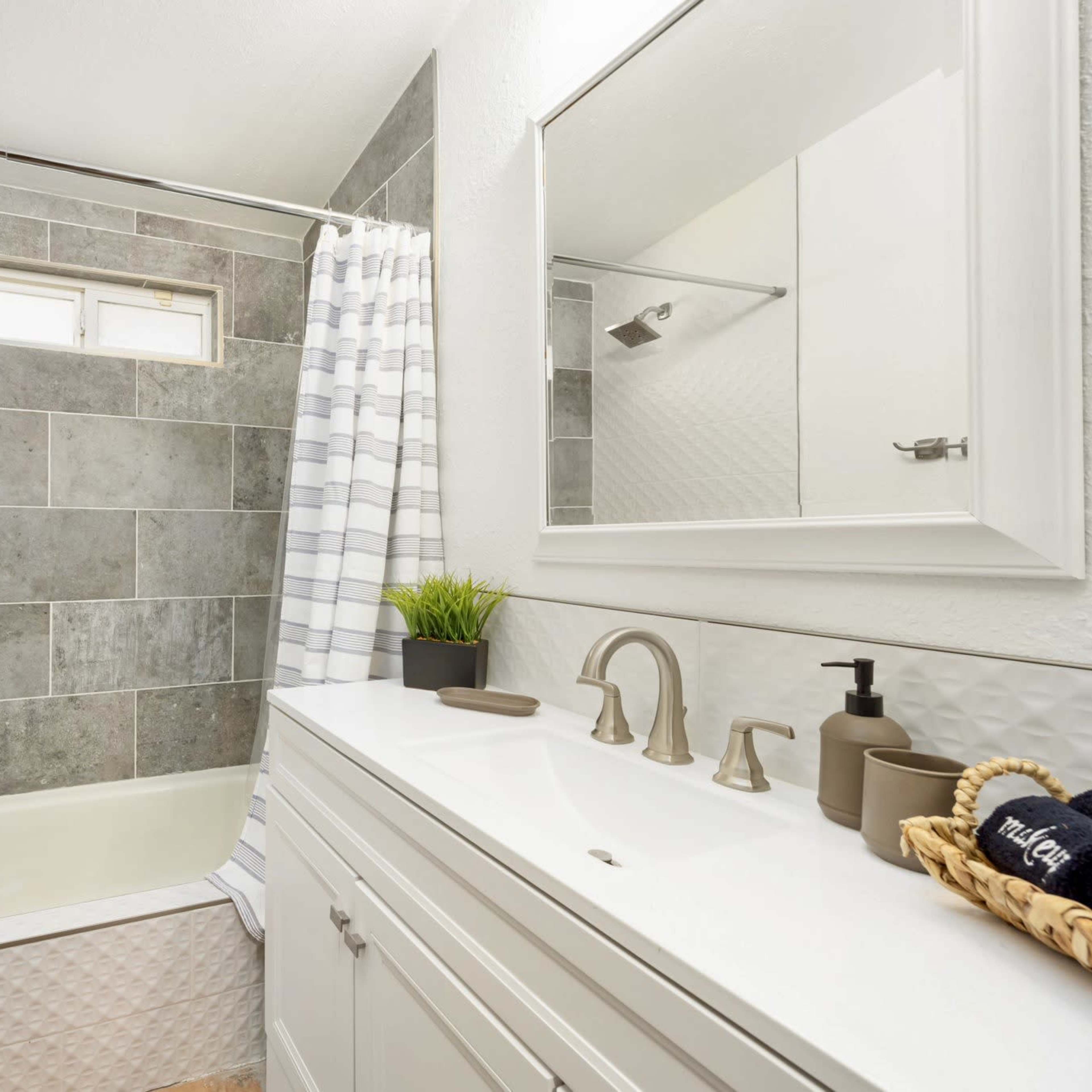 A bathroom featuring a bathtub with a shower curtain, gray tiled walls, a white vanity with a sink, and a mirror above it.