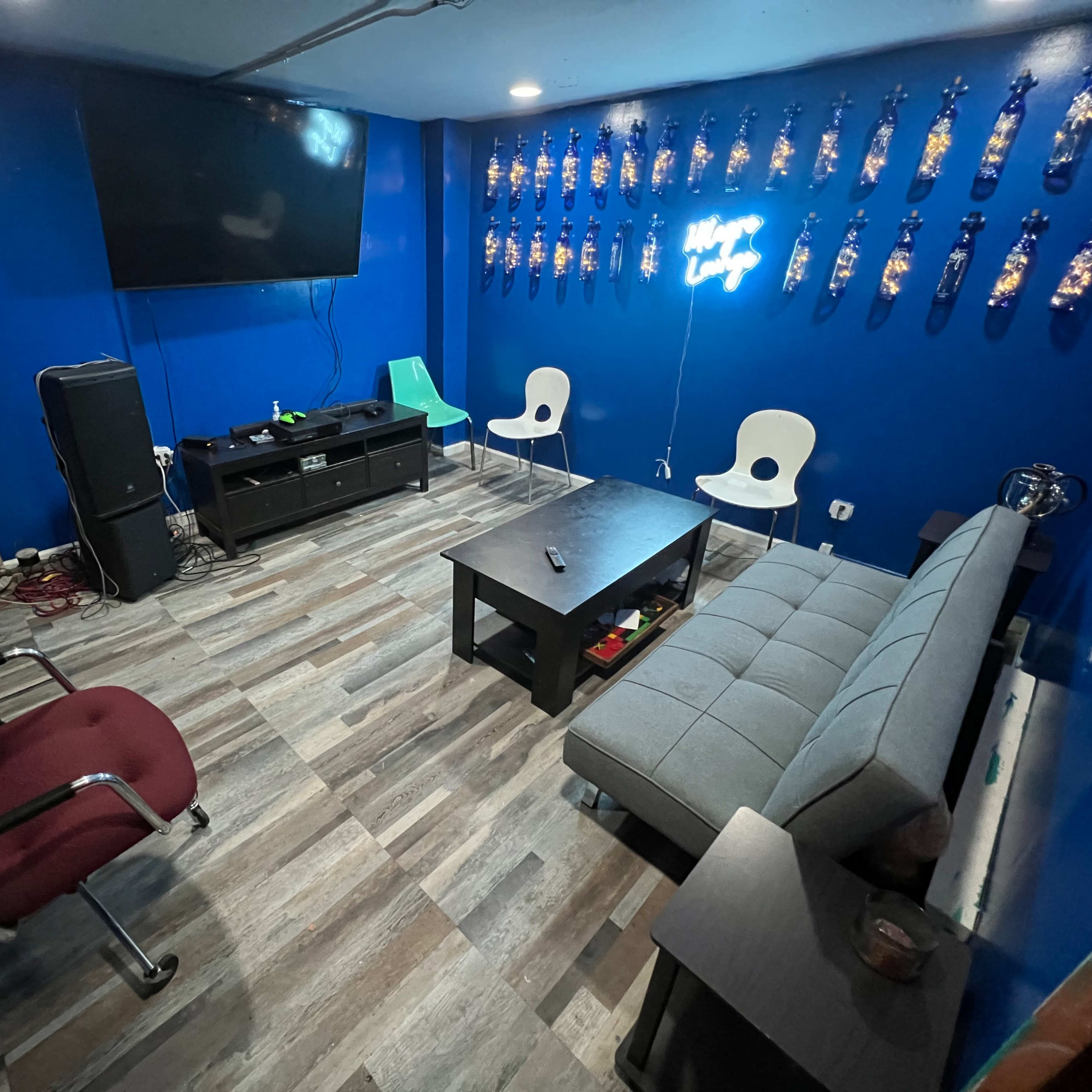 A game room features a large TV mounted on blue walls, a gray couch, a black coffee table, and white chairs, with decorative wall hangings and a few electronic devices scattered around.