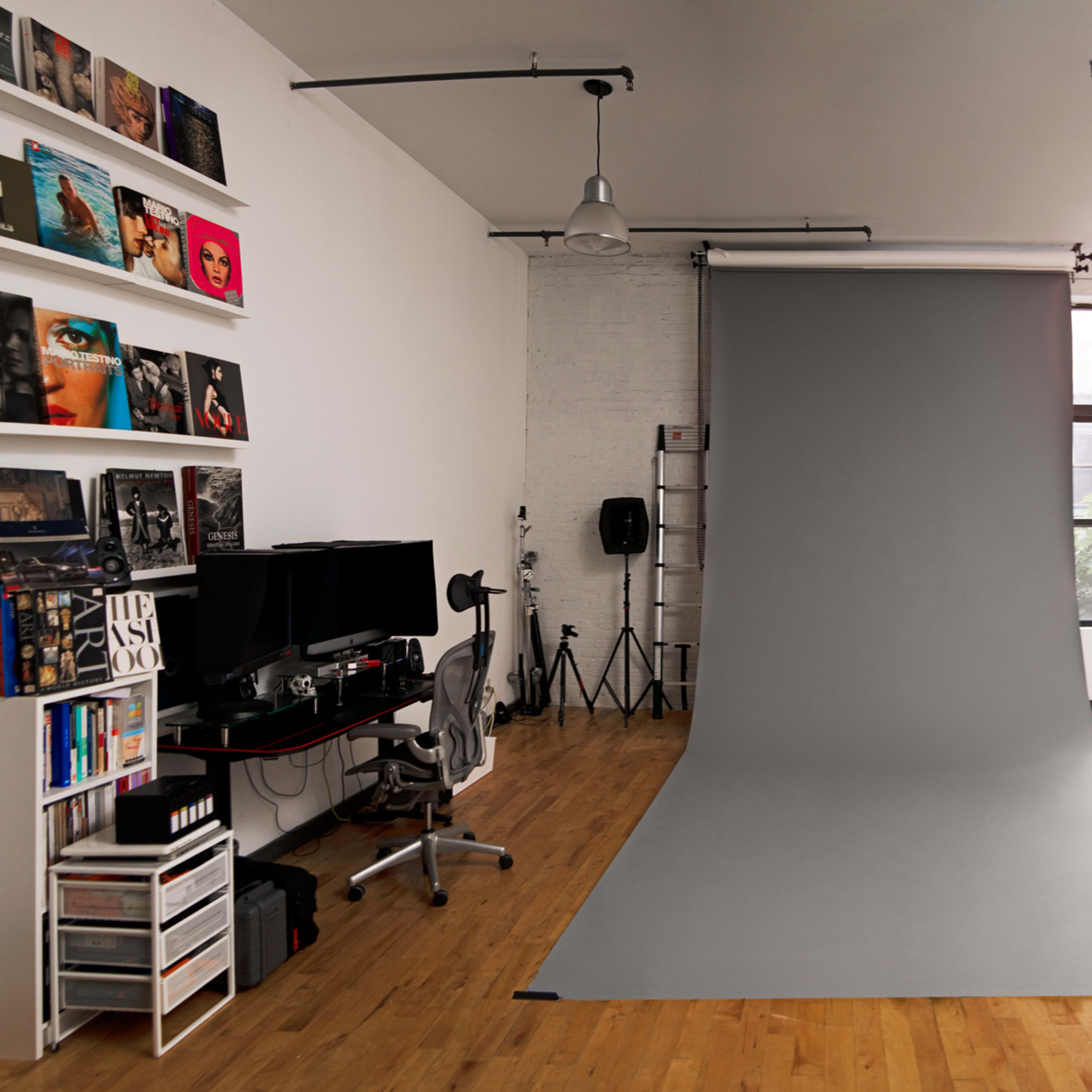 A minimalist studio space with a gray photography backdrop, a desk with dual monitors, and various equipment arranged along the walls.