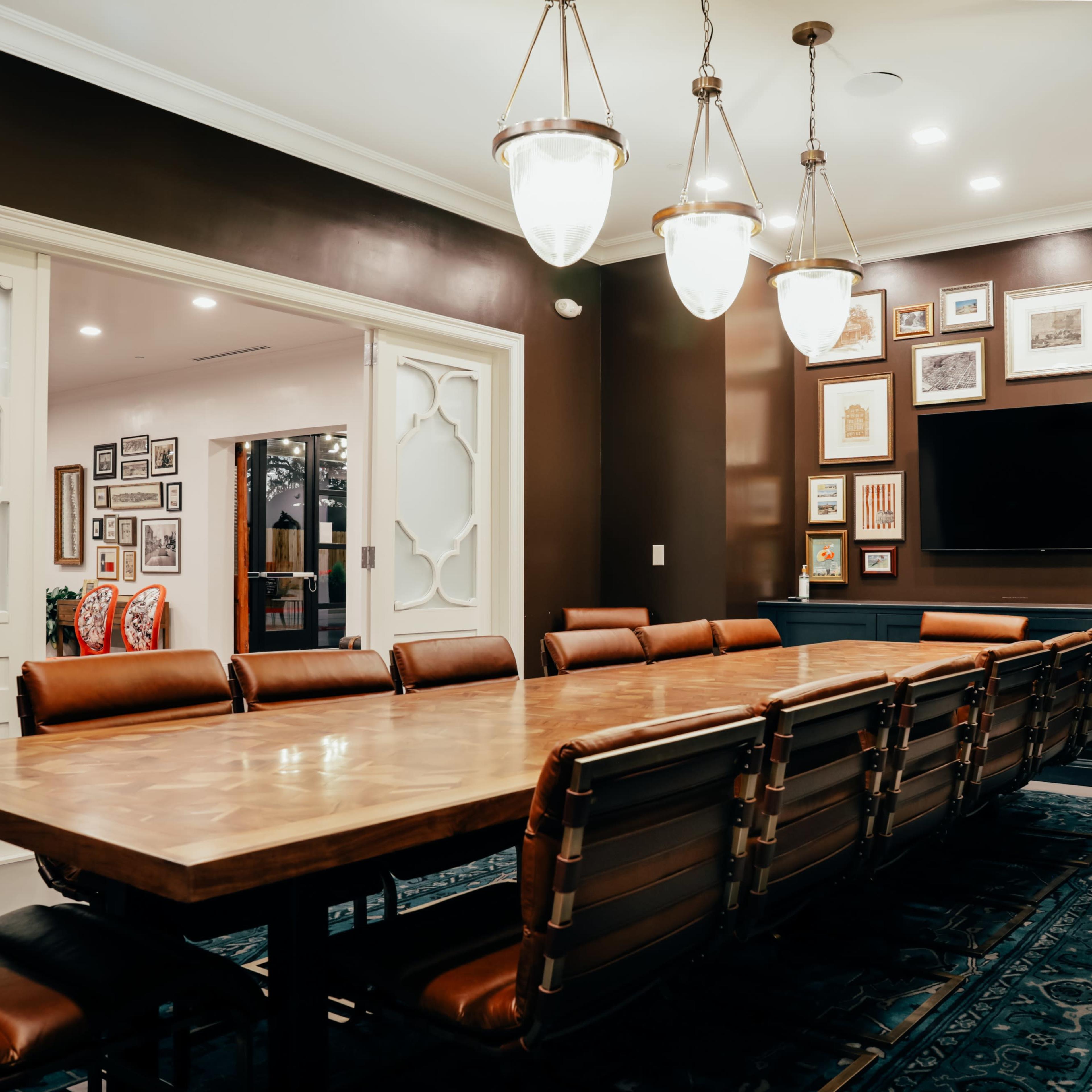 A large conference room features a wooden table surrounded by leather chairs and a wall adorned with framed artwork, with a television mounted above a dark cabinet.