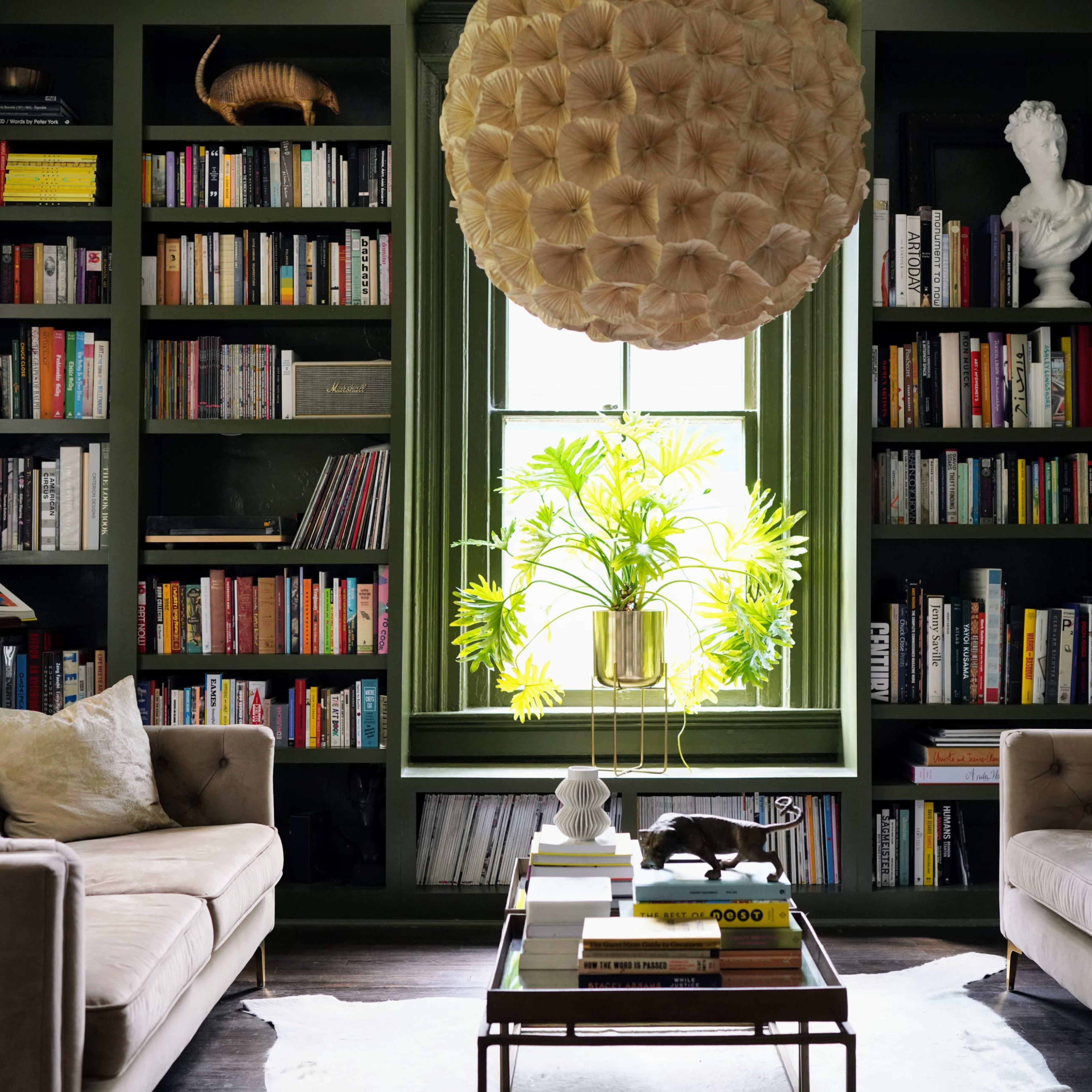 A cozy reading nook features two beige sofas facing a bookshelf filled with books, illuminated by natural light from a large window with a potted plant.