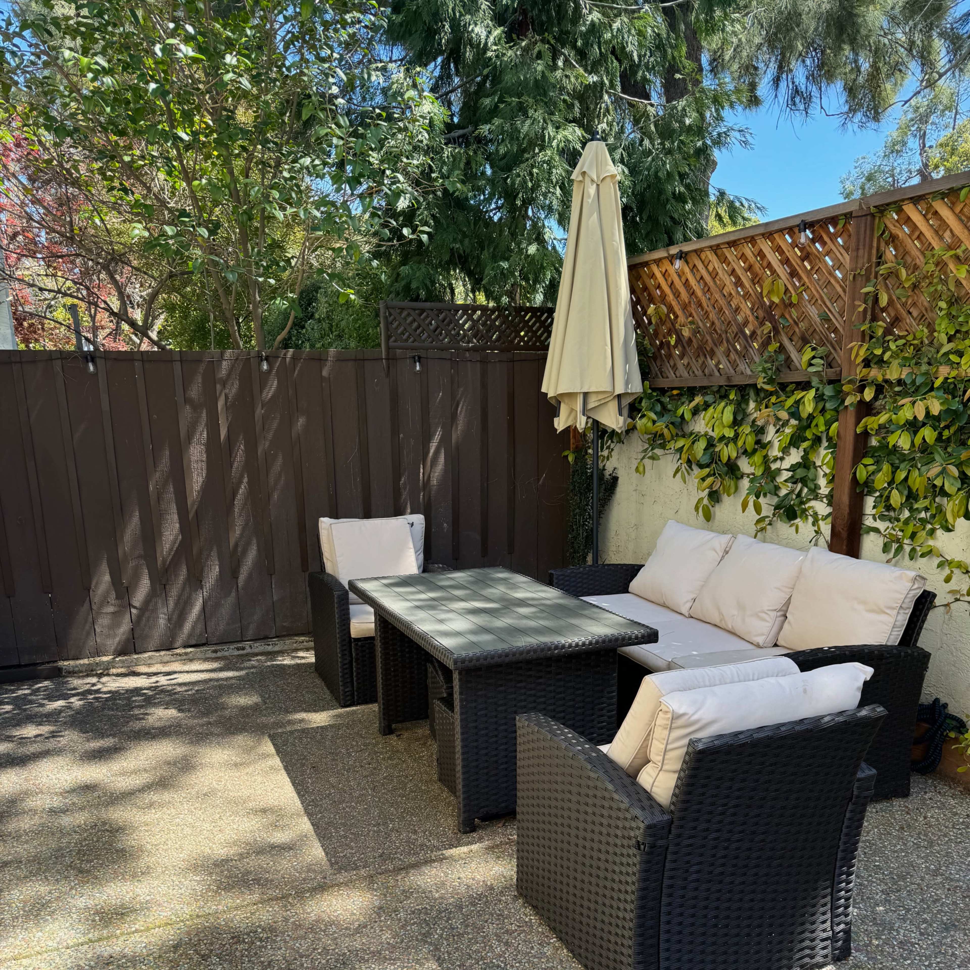 A patio area with a table and four cushioned chairs, surrounded by a wooden fence and lush greenery.