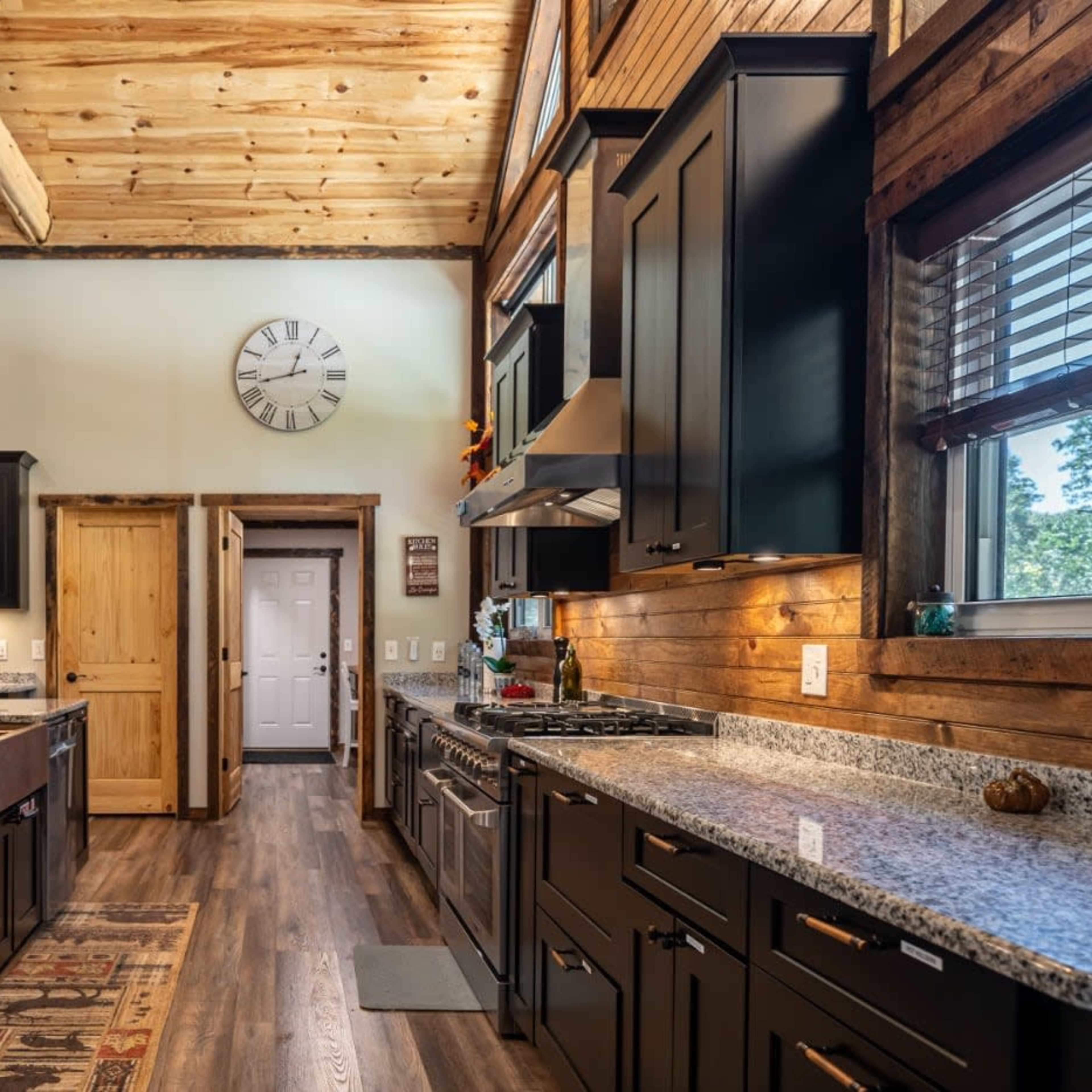 A modern kitchen features dark cabinetry, granite countertops, and wooden beams with a large clock on the wall.