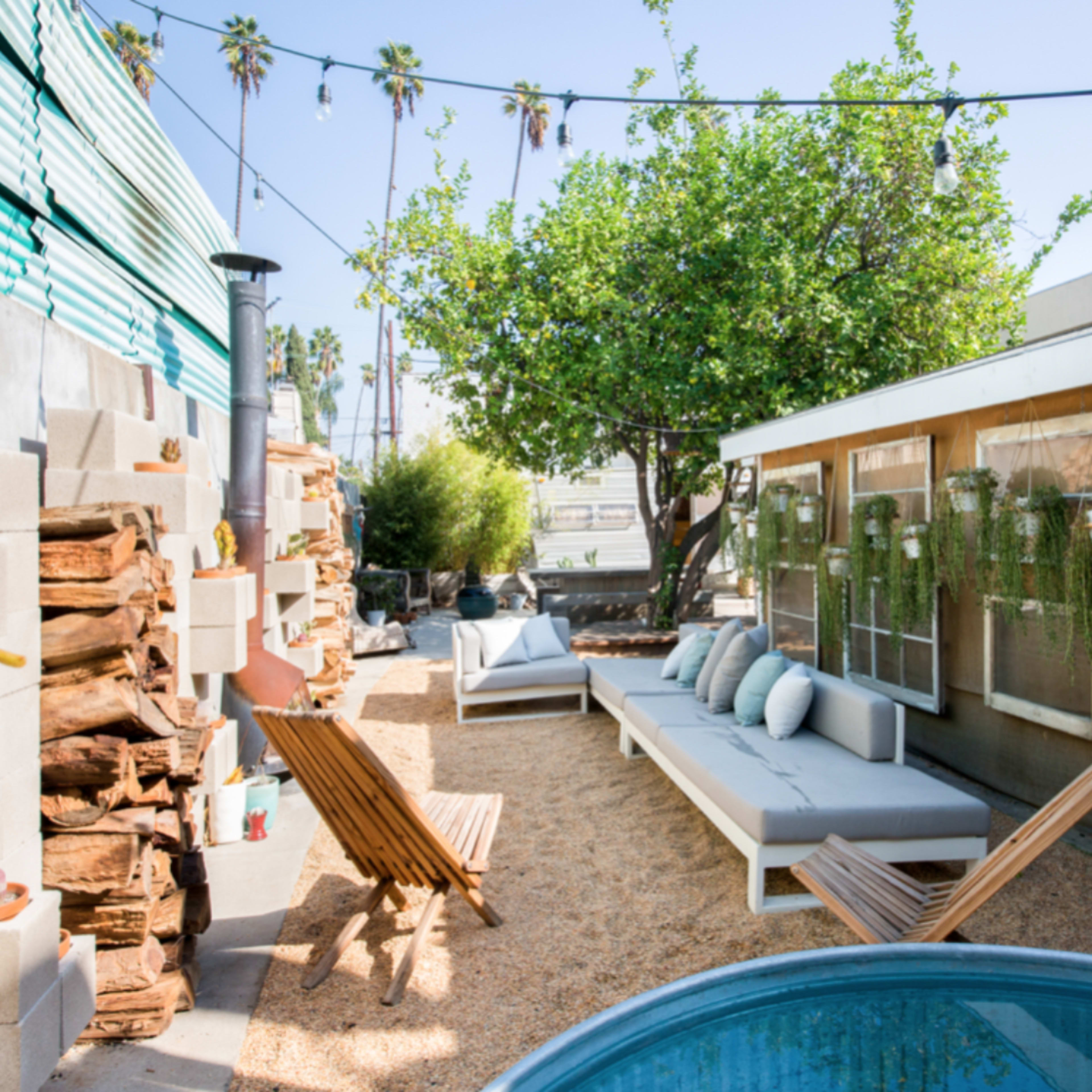 A small outdoor space with seating, a blue pool, green plants, stacked firewood, and a wall featuring decorative plant holders.