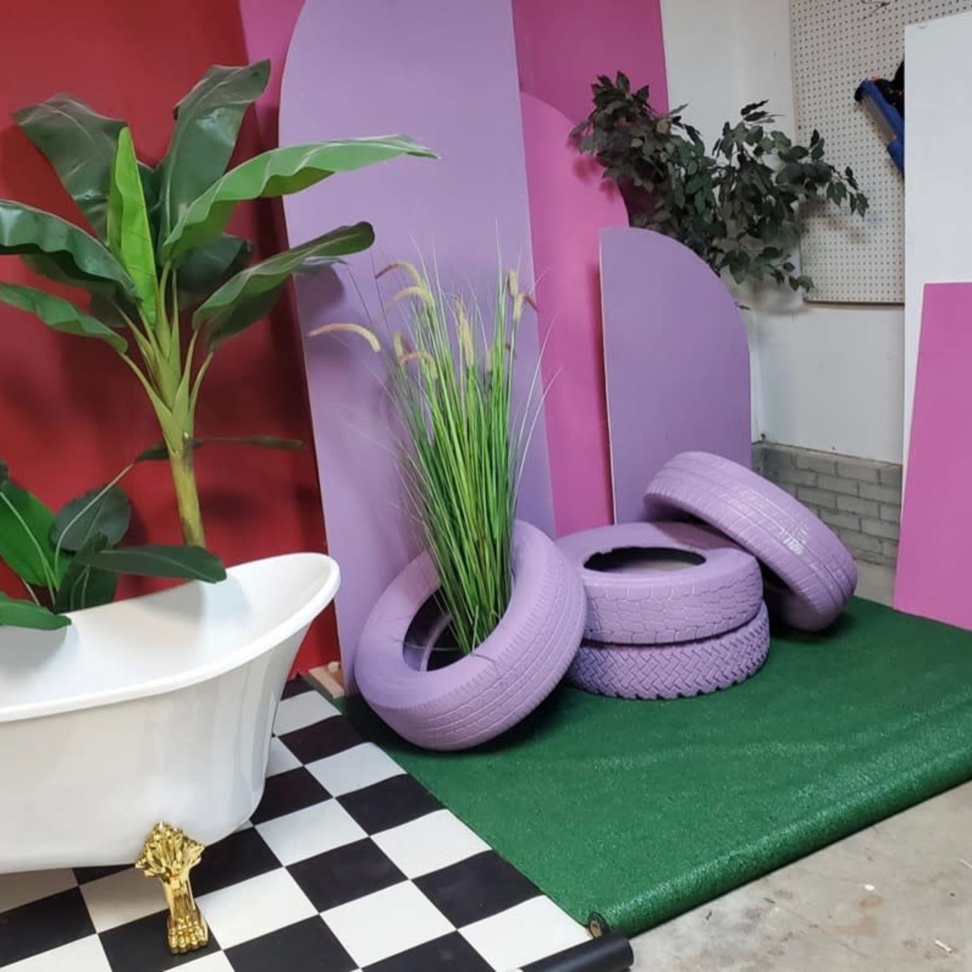 The scene features a white clawfoot bathtub surrounded by decorative plants, purple tire planters, and a checkered black-and-white floor against a backdrop of red and purple panels.