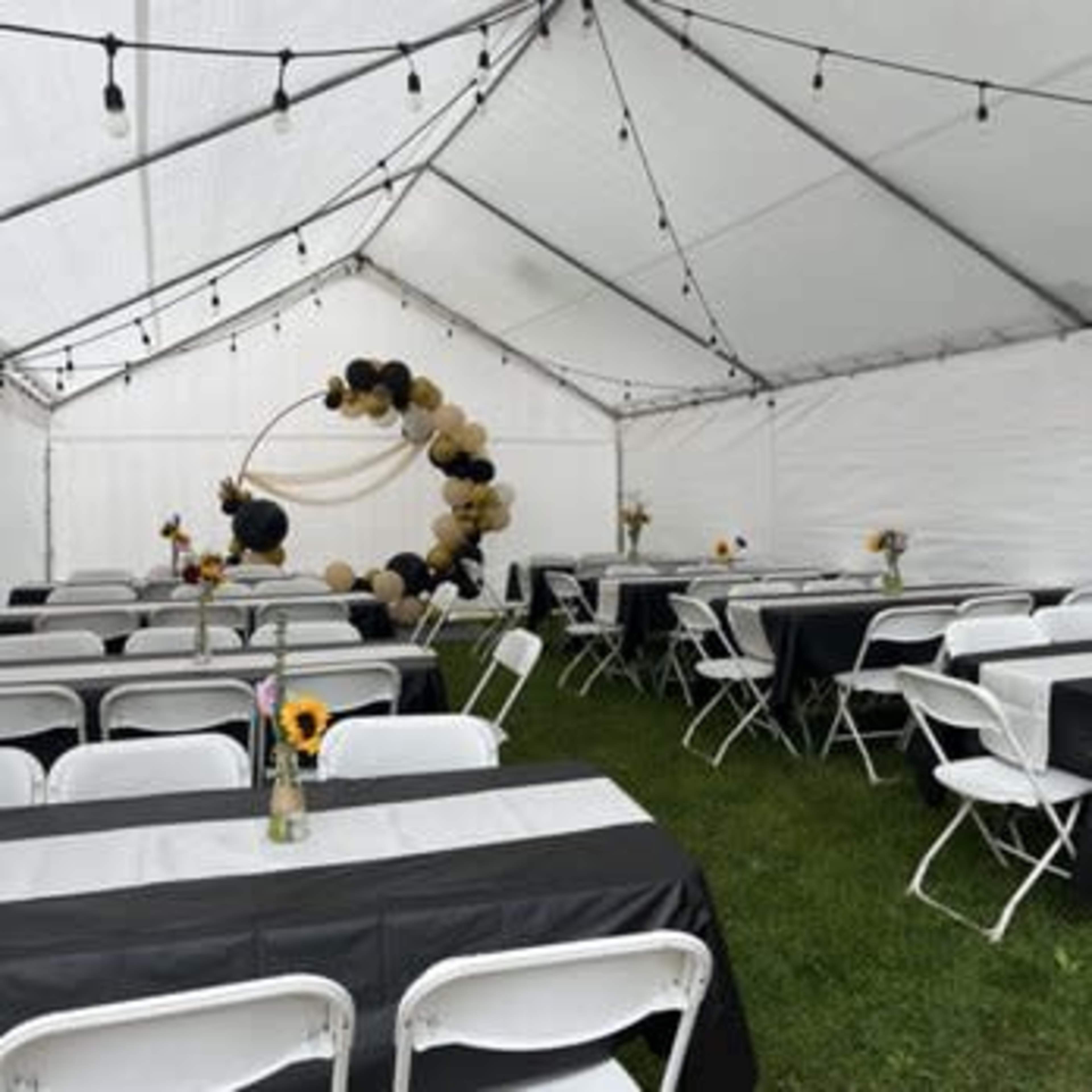 A white tent is set up with rows of tables covered in black and white linens, featuring vases with sunflowers, and a decorative balloon arch in the background.