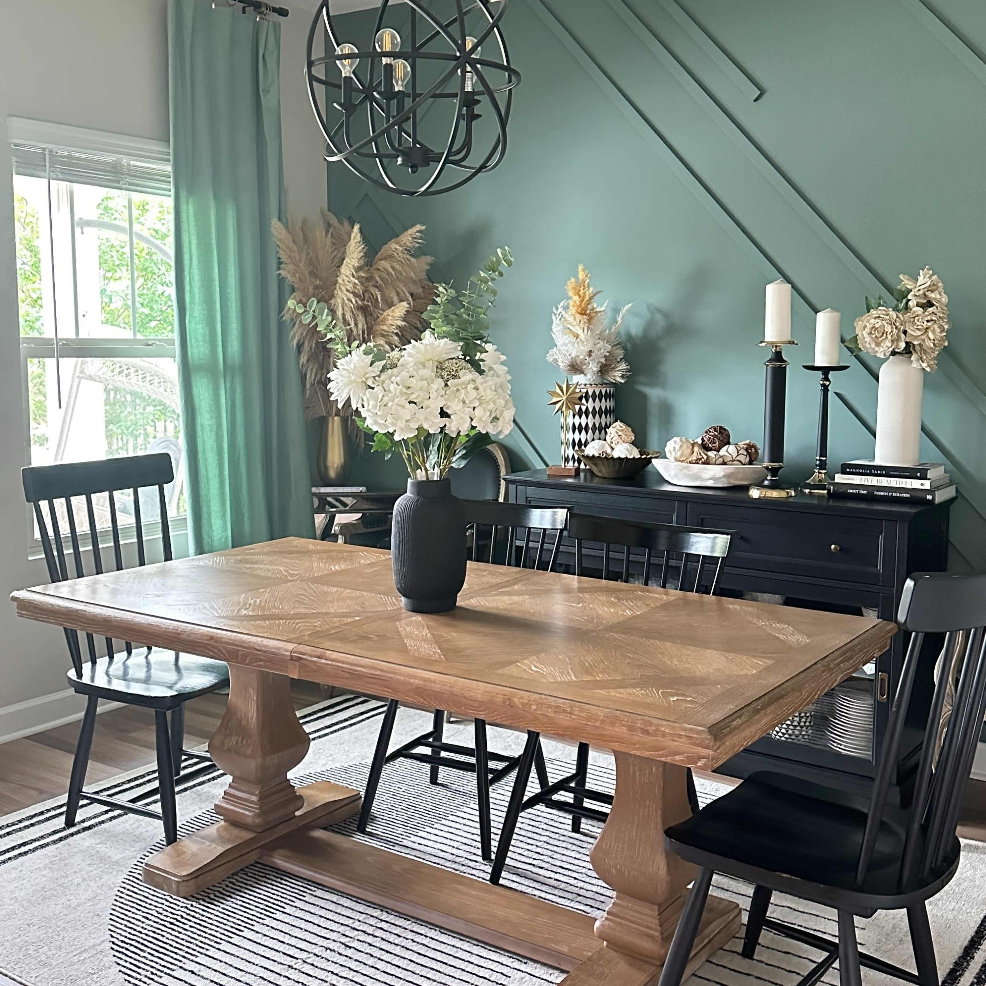 A dining area features a wooden table with black chairs, a vase of flowers, and decorative elements against a green accent wall.