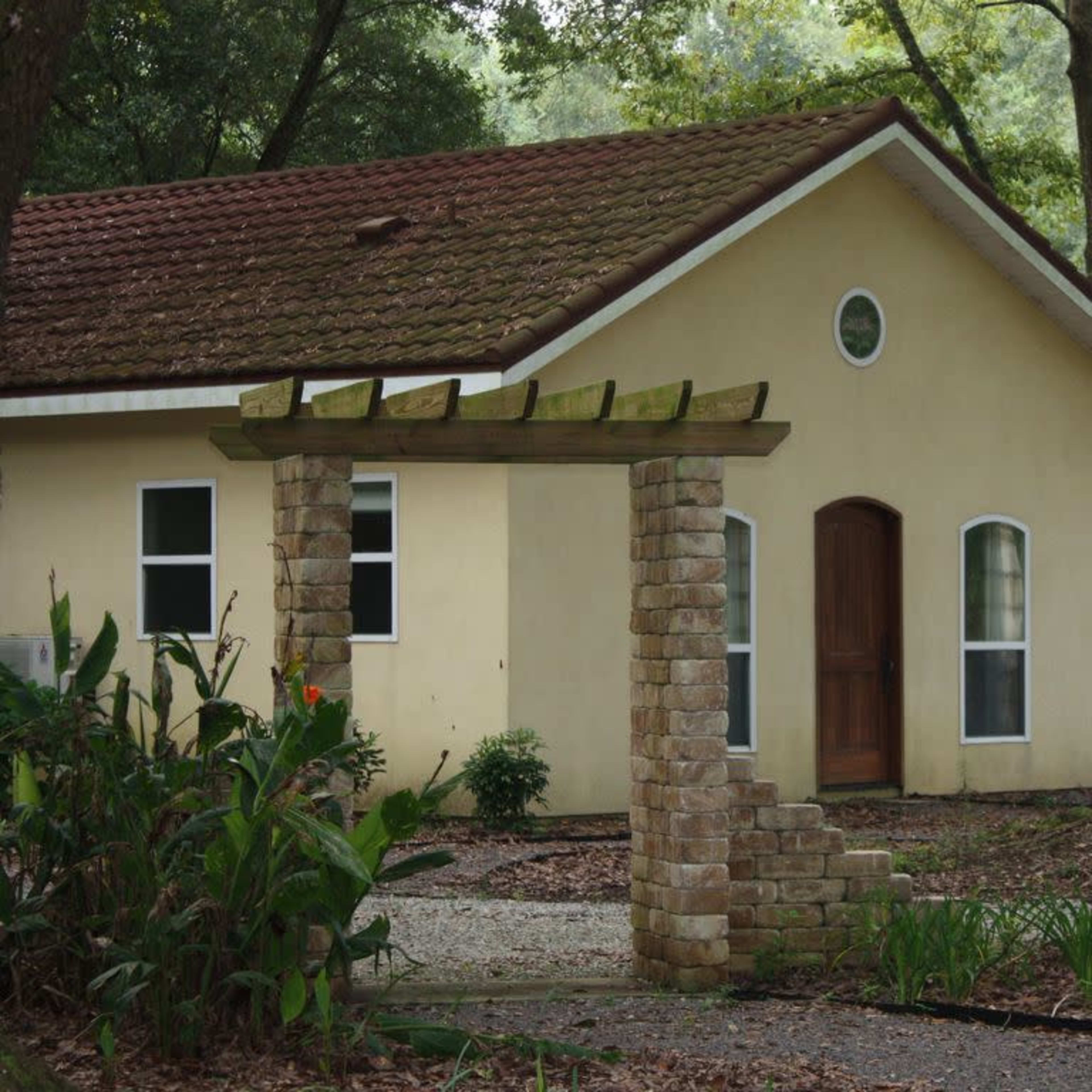 A small house with a brown tiled roof is situated behind a stone archway, surrounded by trees and greenery.