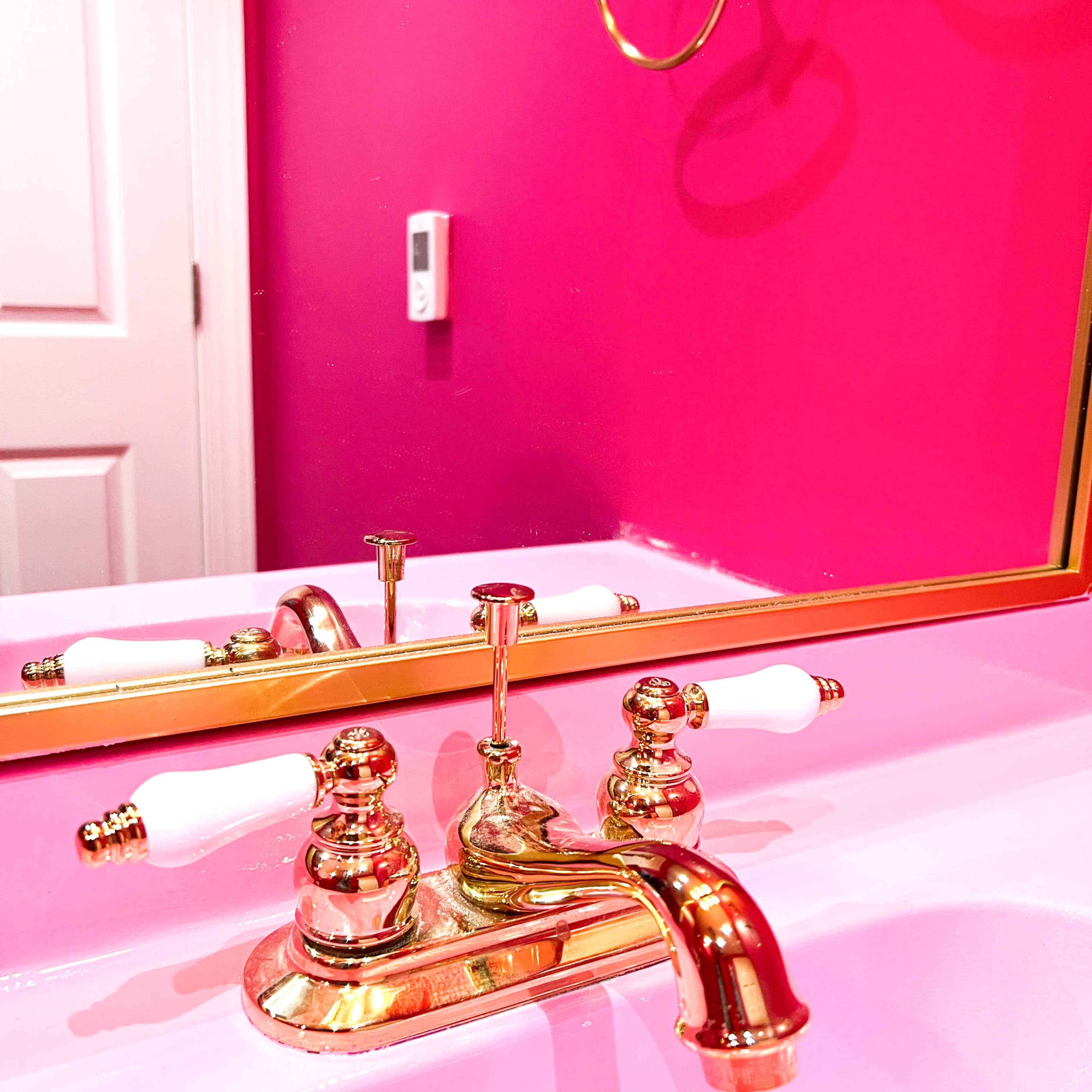 A gold faucet with white handles is reflected in a pink wall mirror above a pink sink.