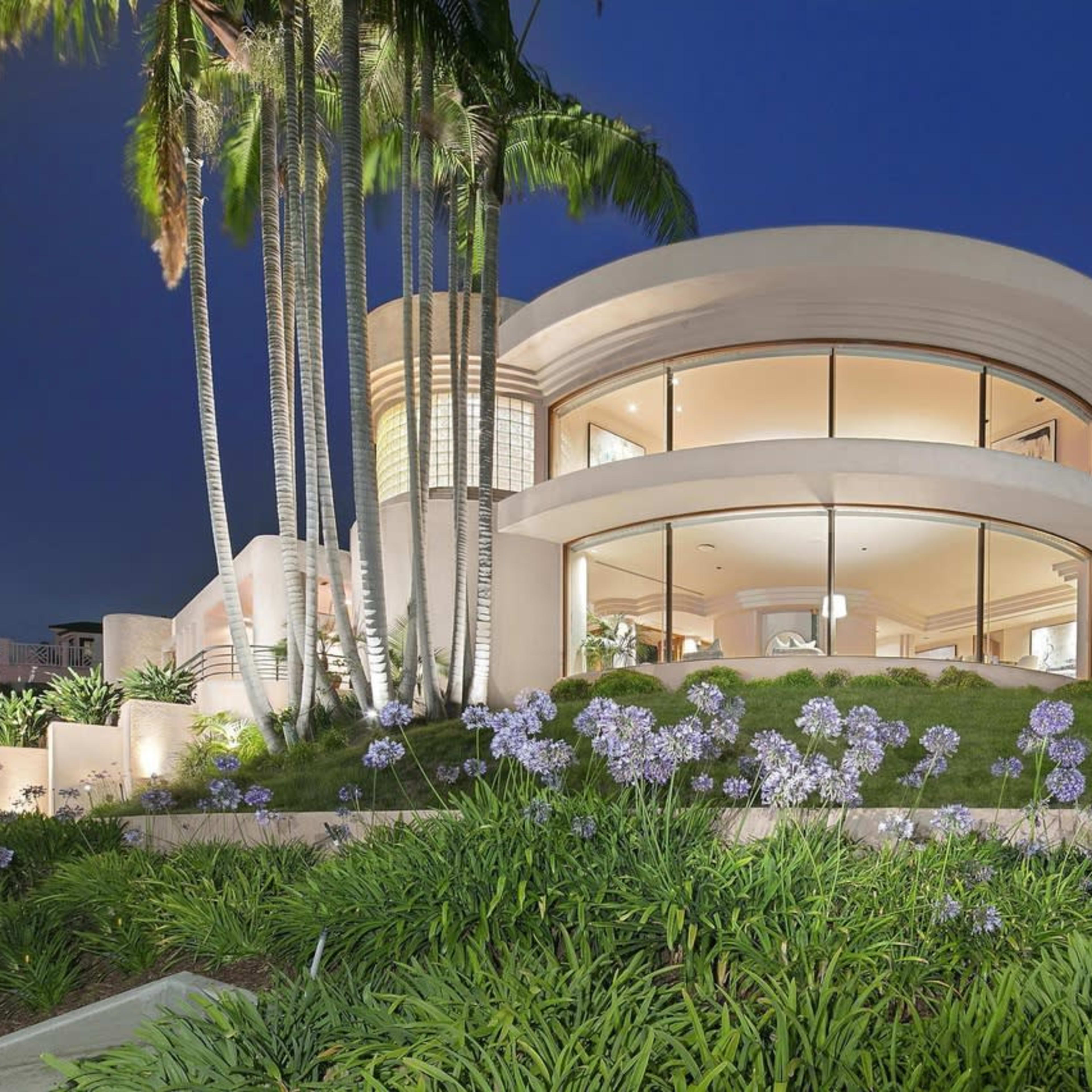 A modern, circular house with large windows, surrounded by manicured gardens and palm trees at dusk.