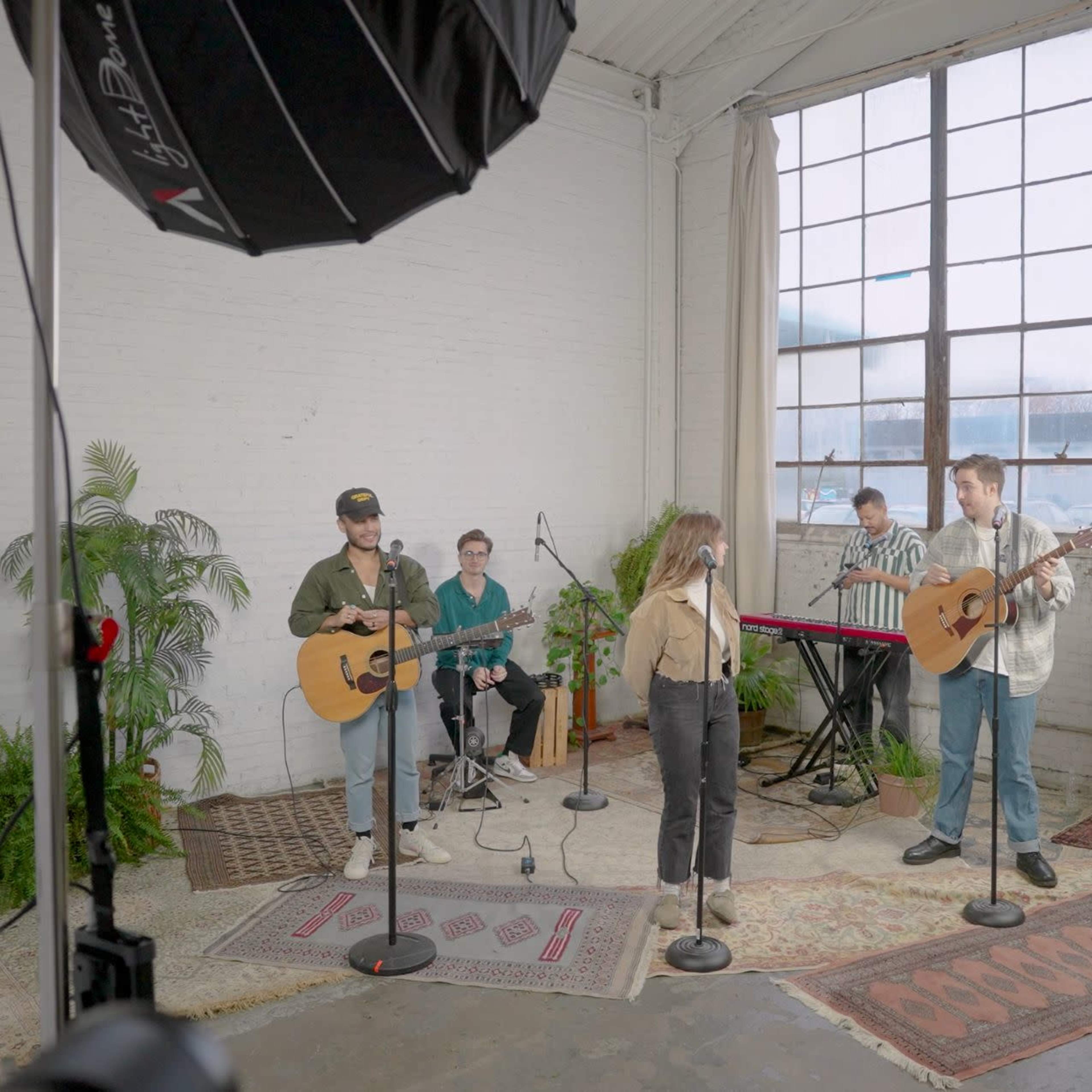A group of musicians is performing with guitars and keyboards in a studio space featuring large windows and potted plants.