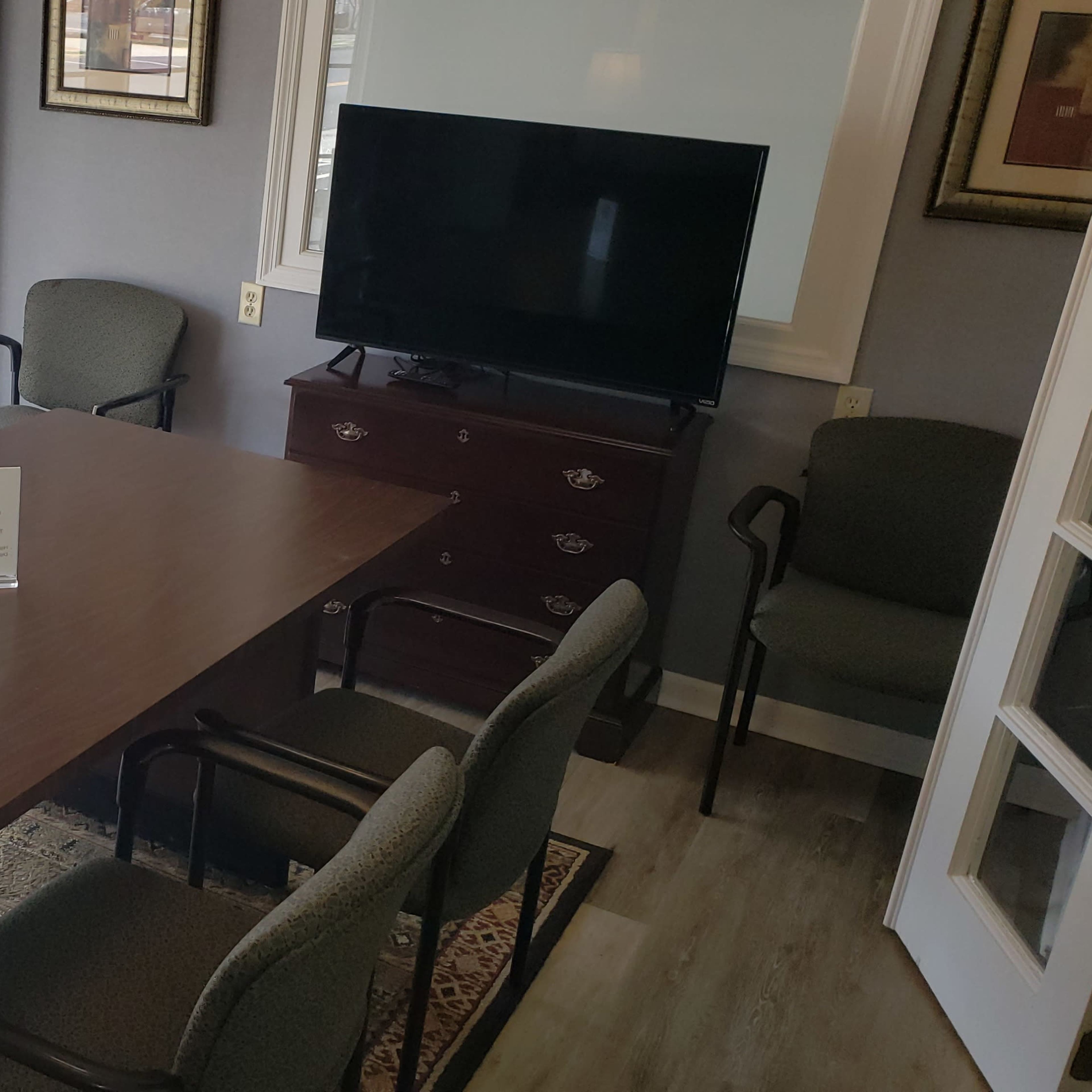 A conference room features a large wooden table surrounded by six chairs, with a television mounted on the wall and large windows allowing natural light.