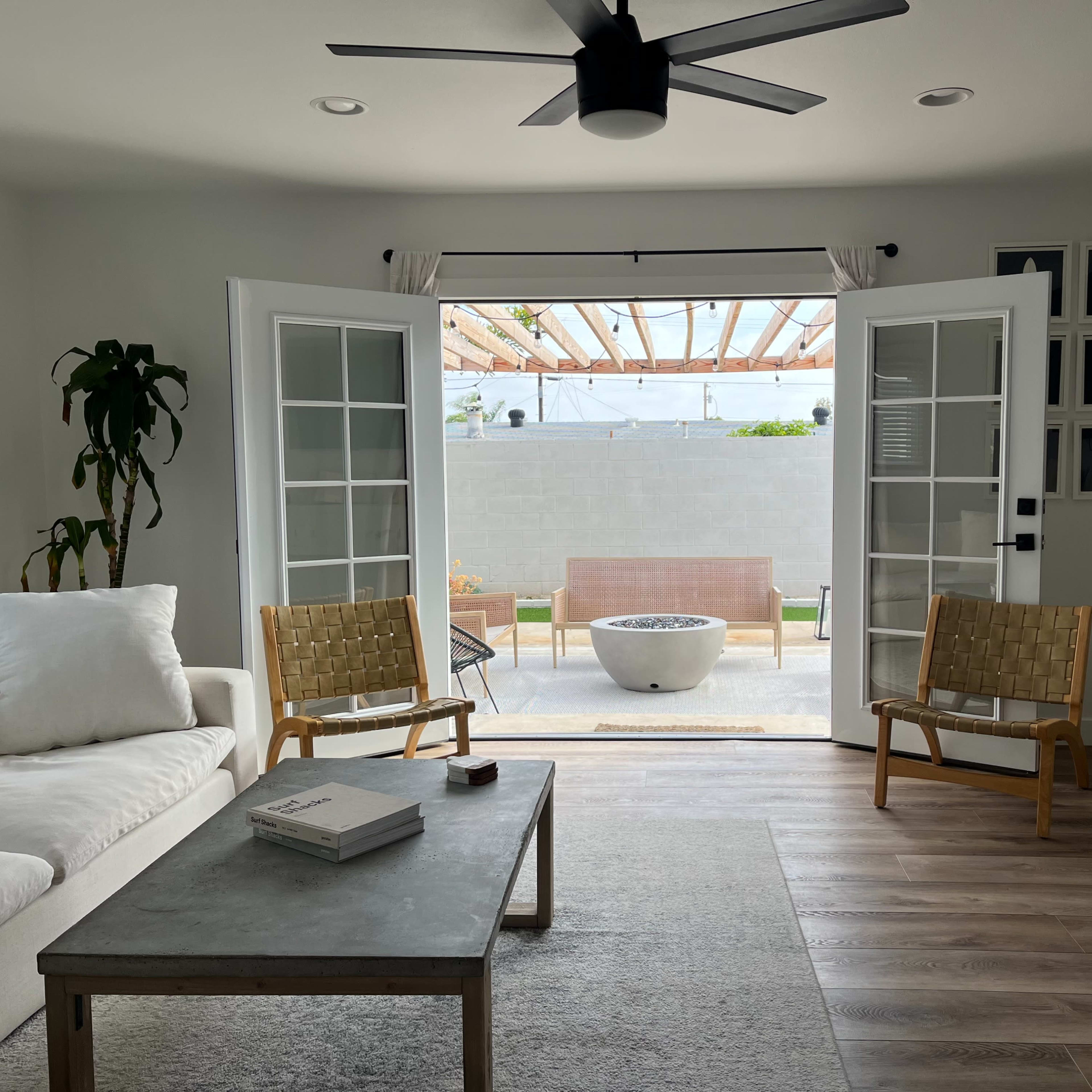 A bright living room features a white sofa, two wooden chairs, and French doors leading to a patio with a stone fire pit and outdoor seating.