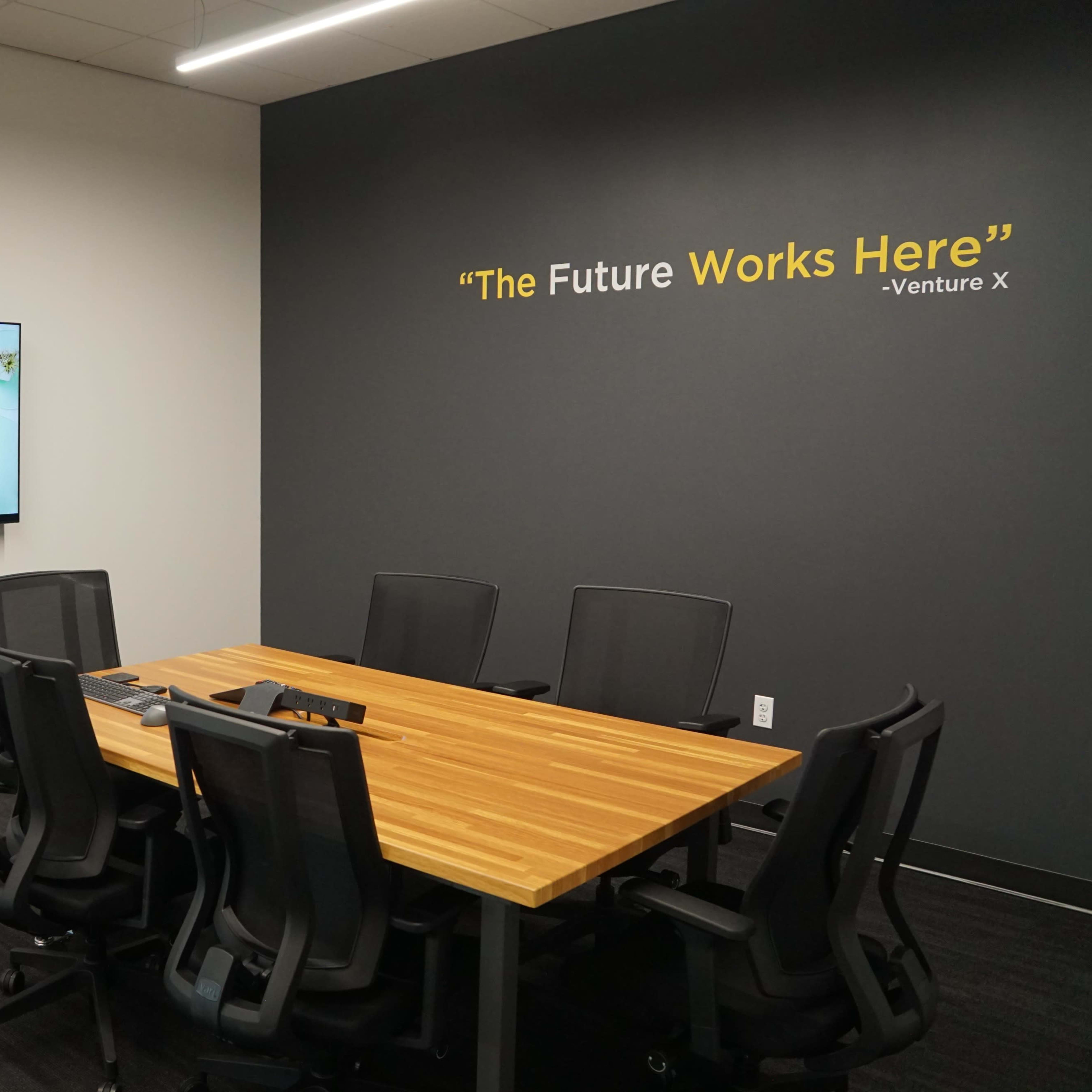 A modern conference room features a large wooden table surrounded by black chairs, with a wall-mounted screen displaying a video conference setup.