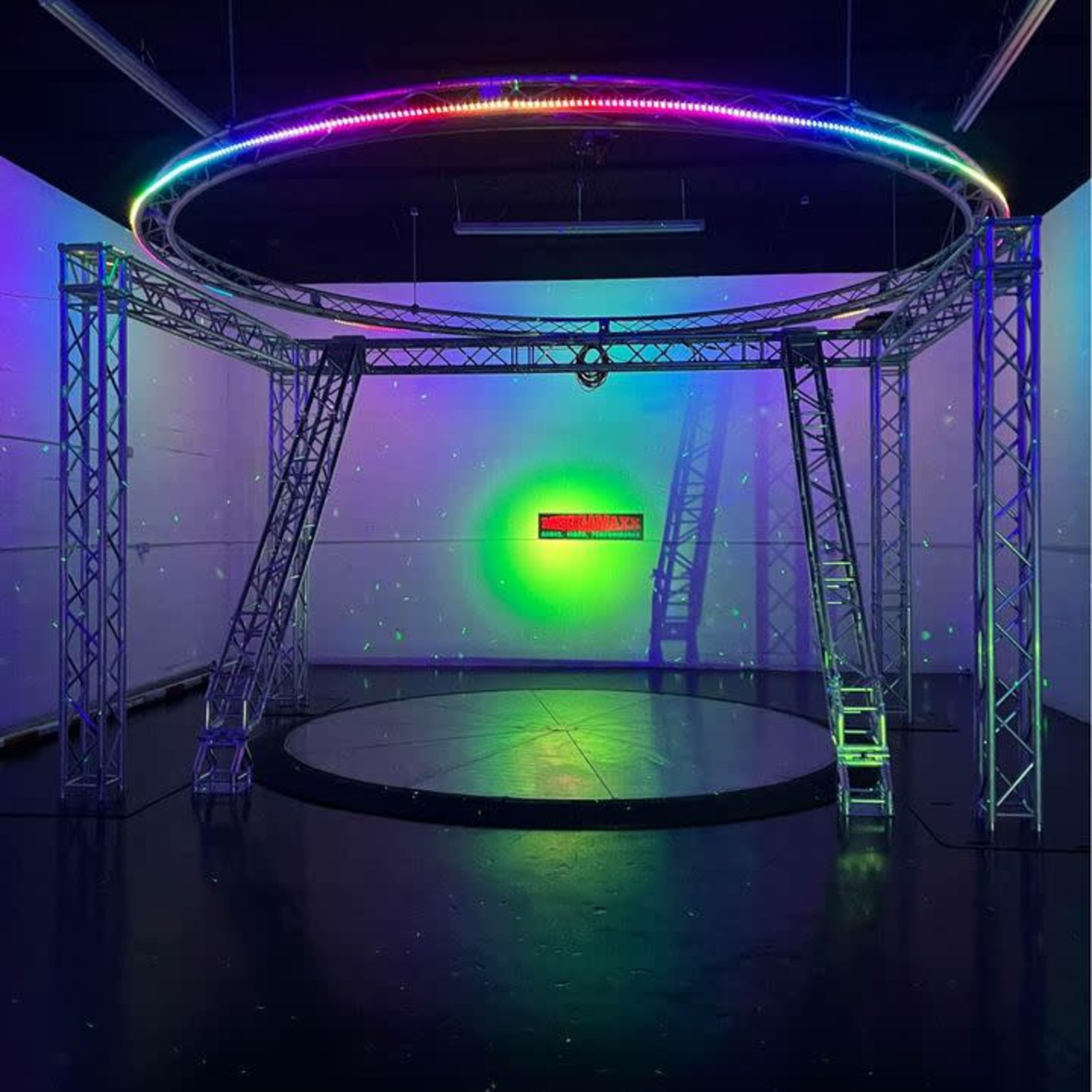 A circular stage structure with bright, multicolored lights and a dark floor in a dimly lit space.
