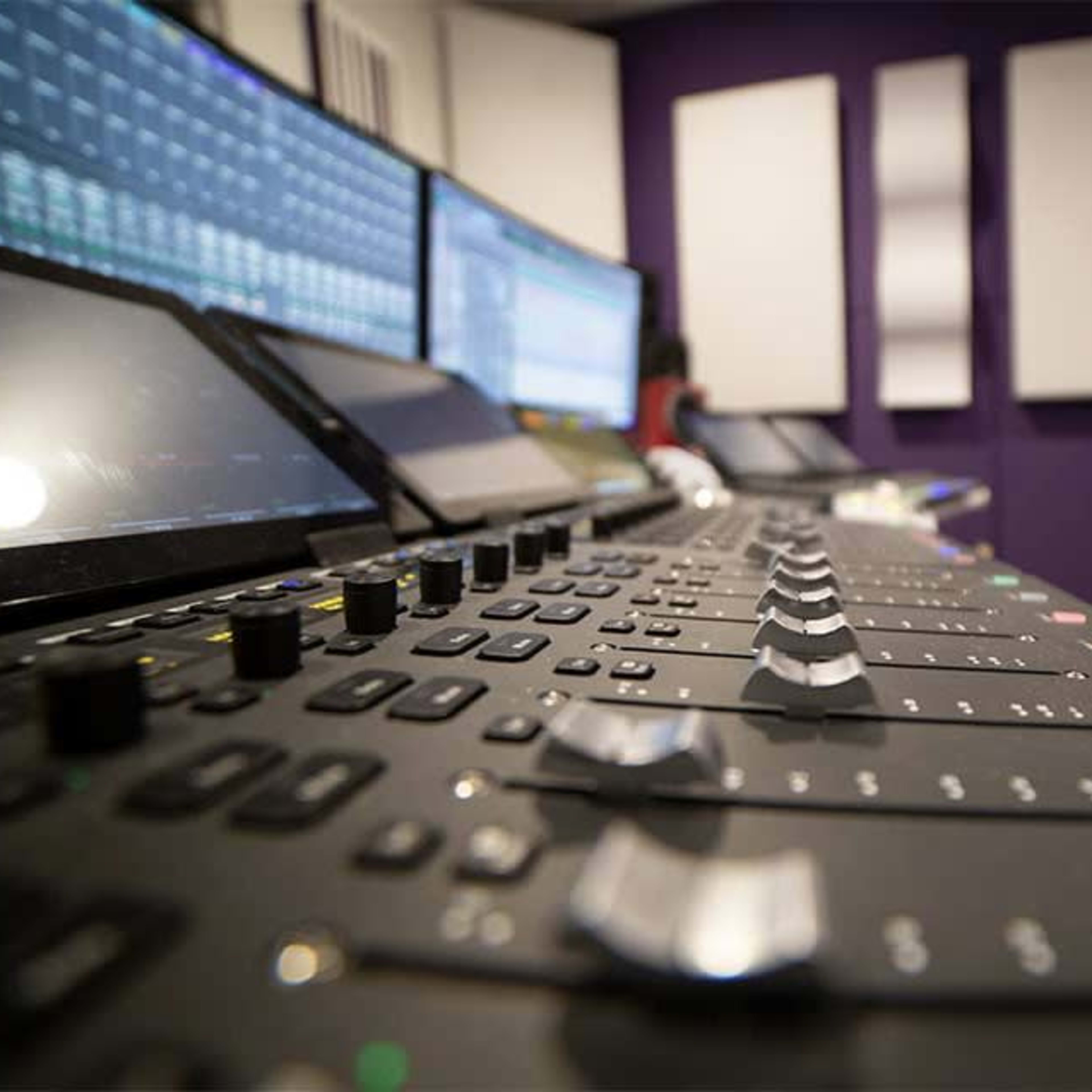 A close-up view of a sound mixing console with multiple faders, set in a recording studio featuring purple walls and screens displaying audio tracks.