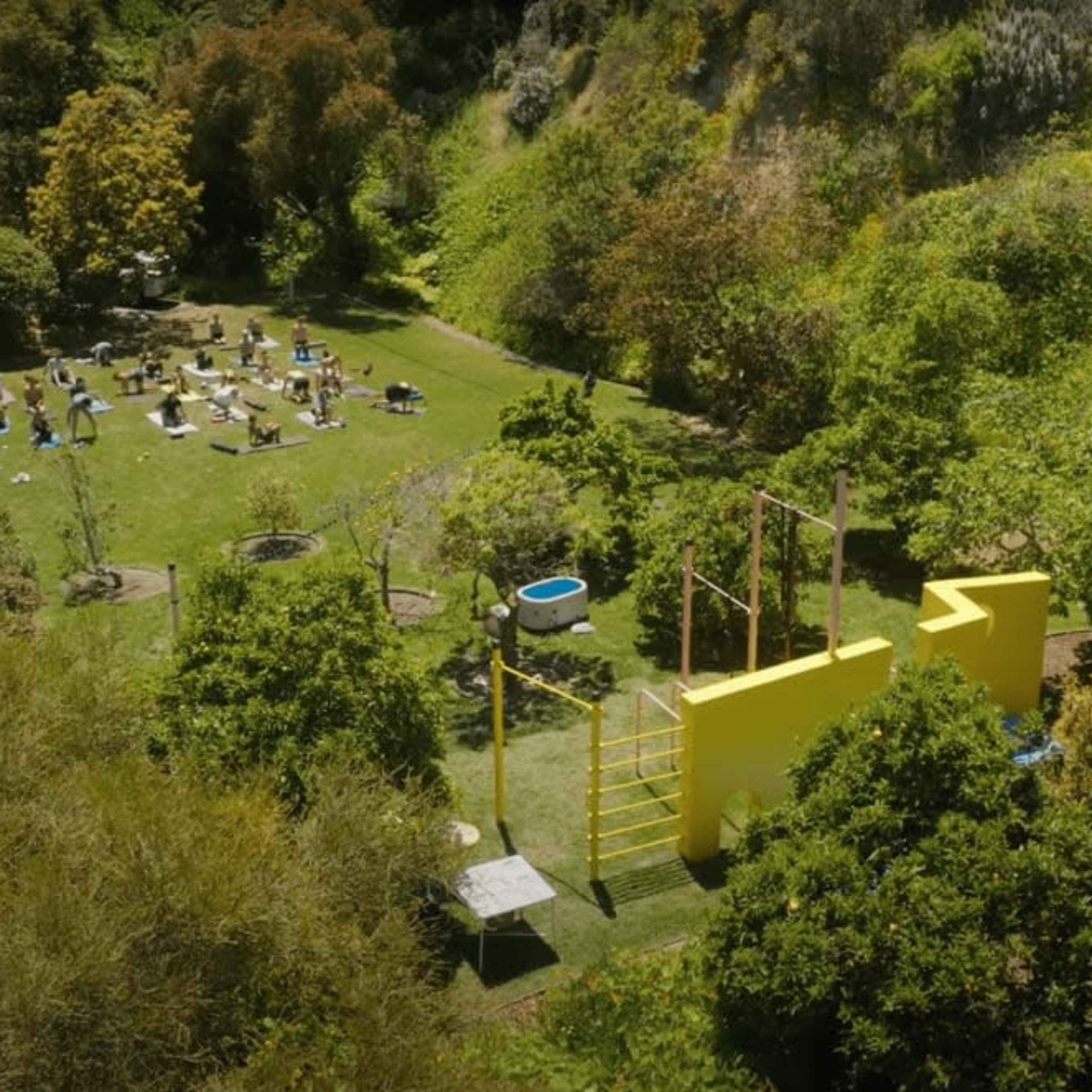 A lush, green outdoor area with various people engaged in activities, surrounded by trees and marked by a large yellow structure.