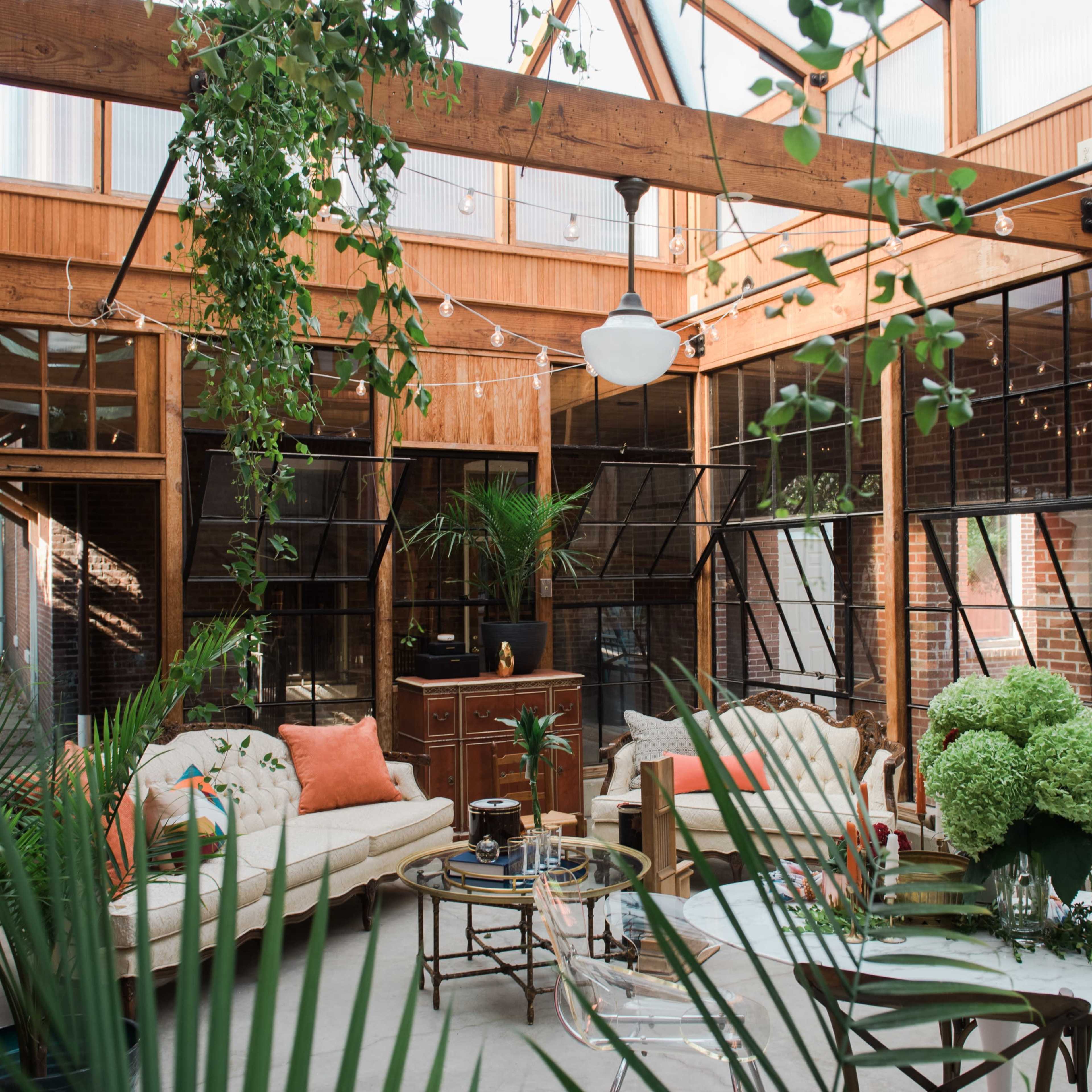 A bright and open indoor space featuring a living area with a sofa, plants, and a round dining table under a glass roof.