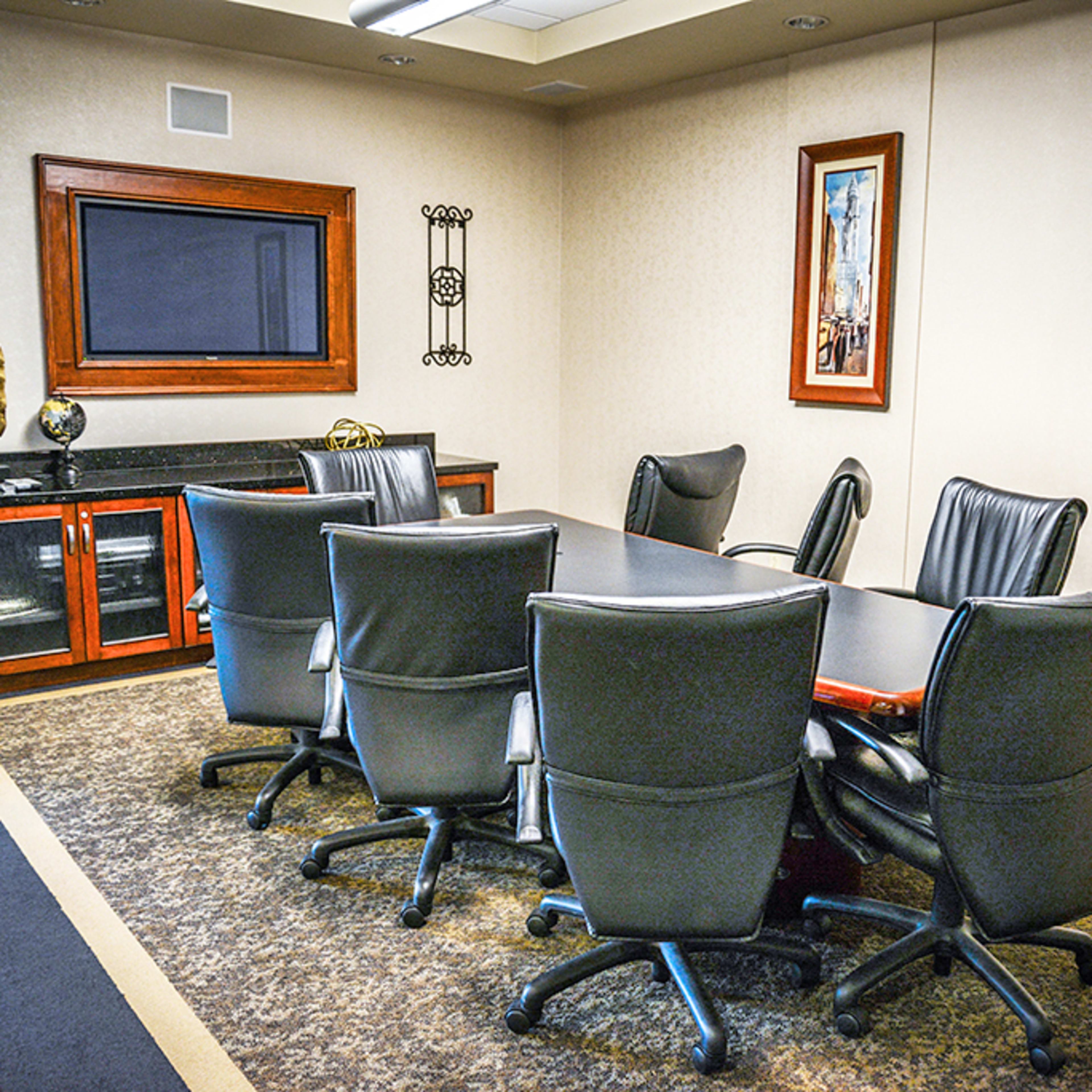 A conference room with a large rectangular table surrounded by black office chairs, two framed pictures on the walls, and a wall-mounted television.