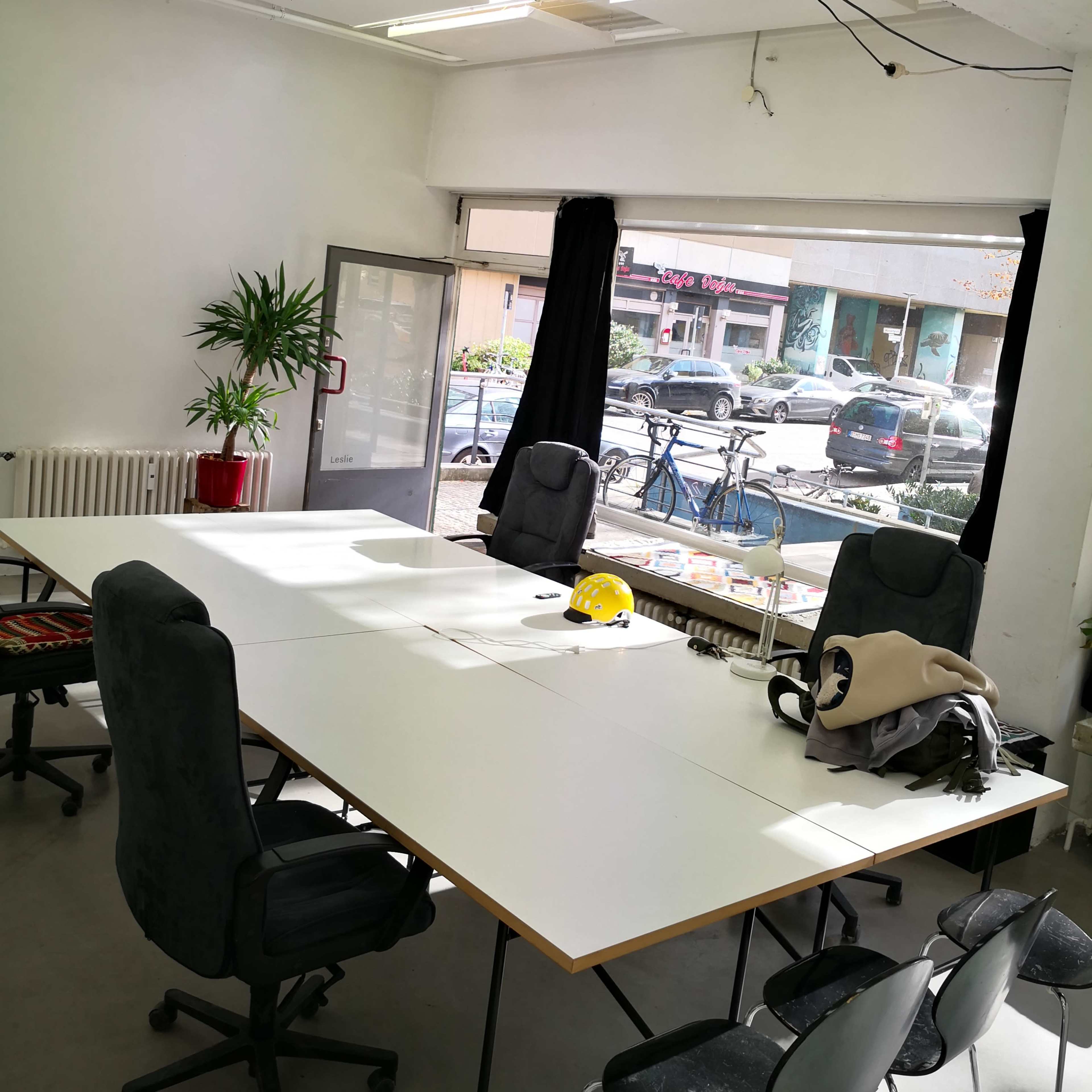 A brightly lit meeting room features a large table surrounded by chairs, with a projector mounted on the ceiling and a window showing street views outside.
