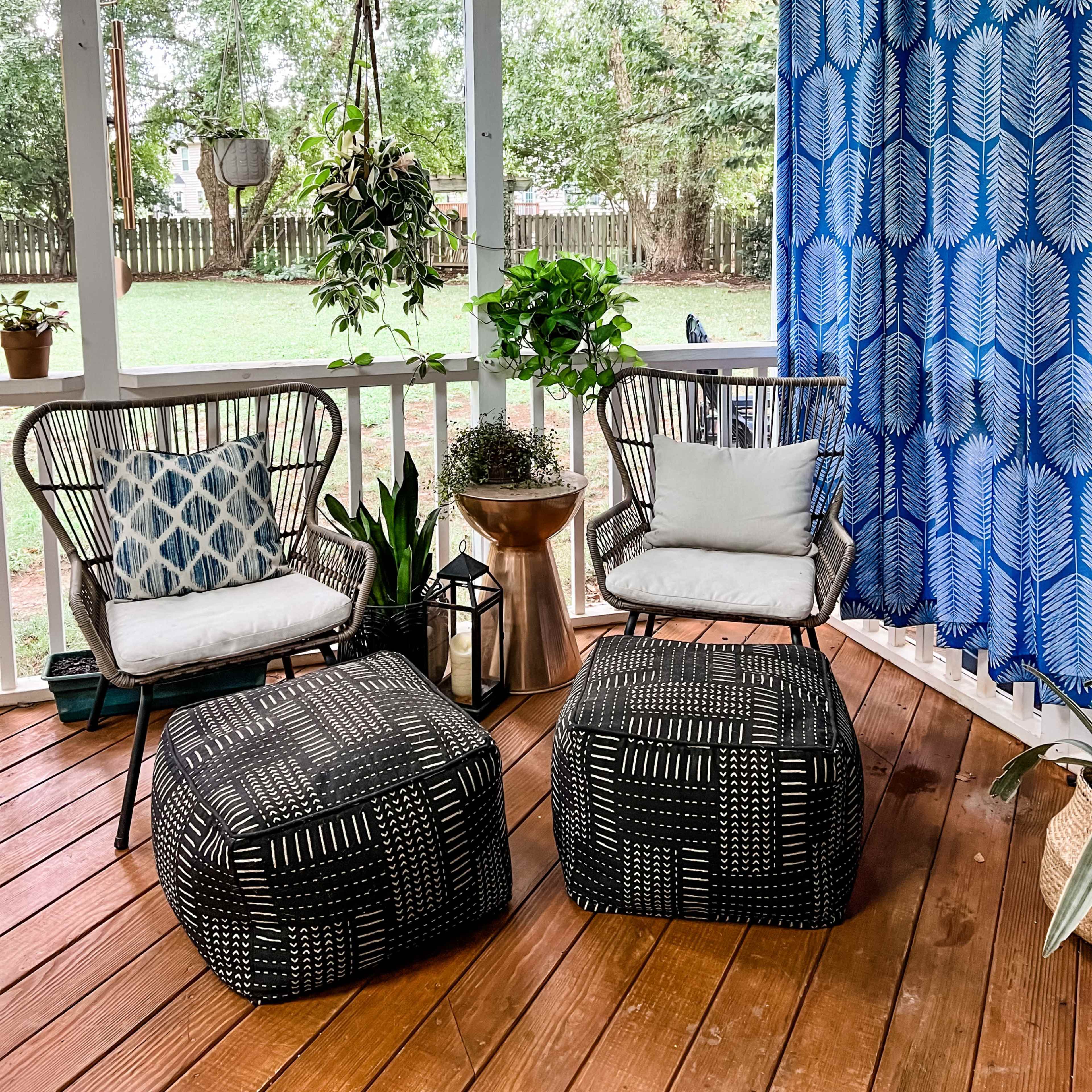A covered outdoor space with two black patterned ottomans, two chairs with cushions, a small table, and plants arranged on a wooden deck.