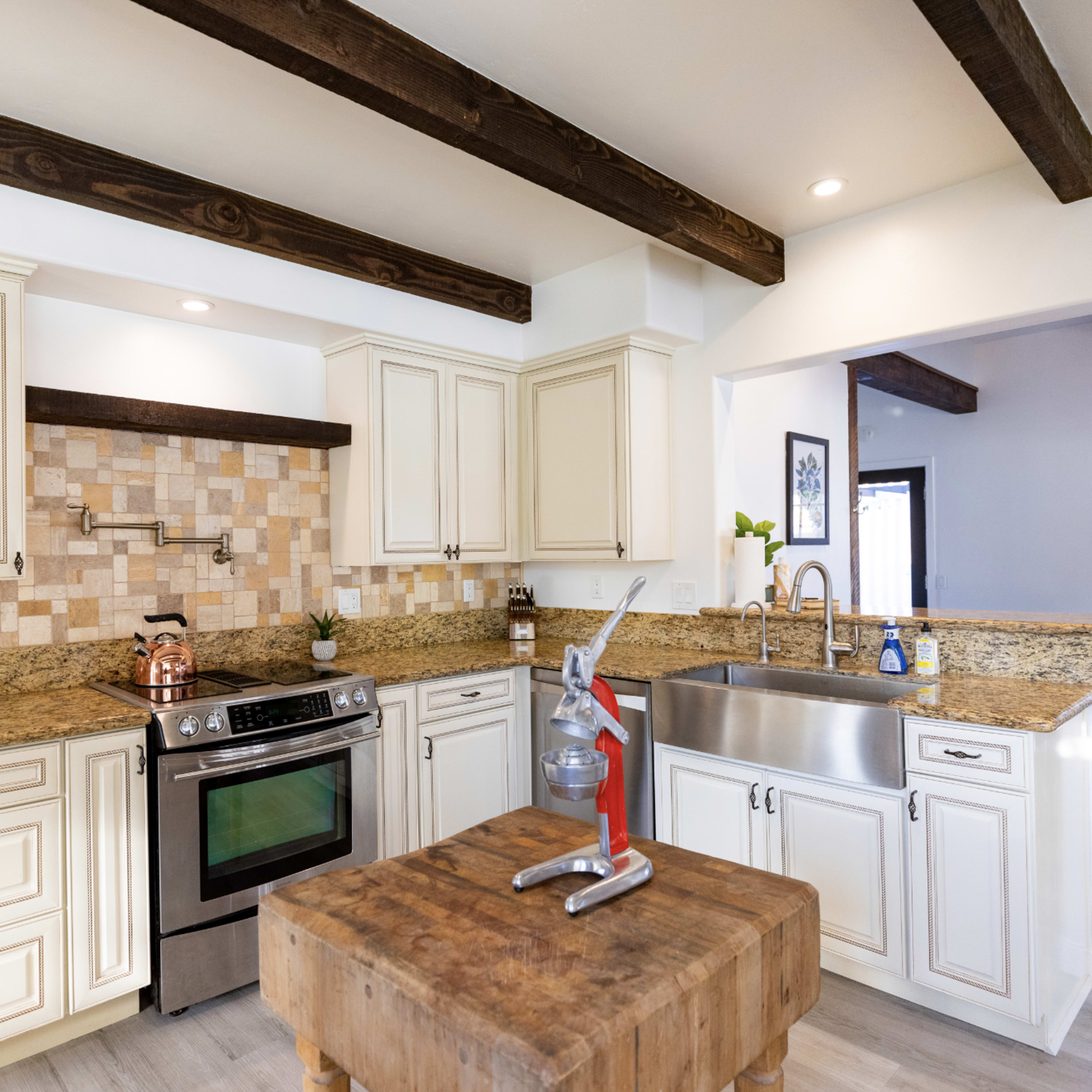 The kitchen features cream-colored cabinets, a stone backsplash, a stainless steel sink, and wooden beams across the ceiling.