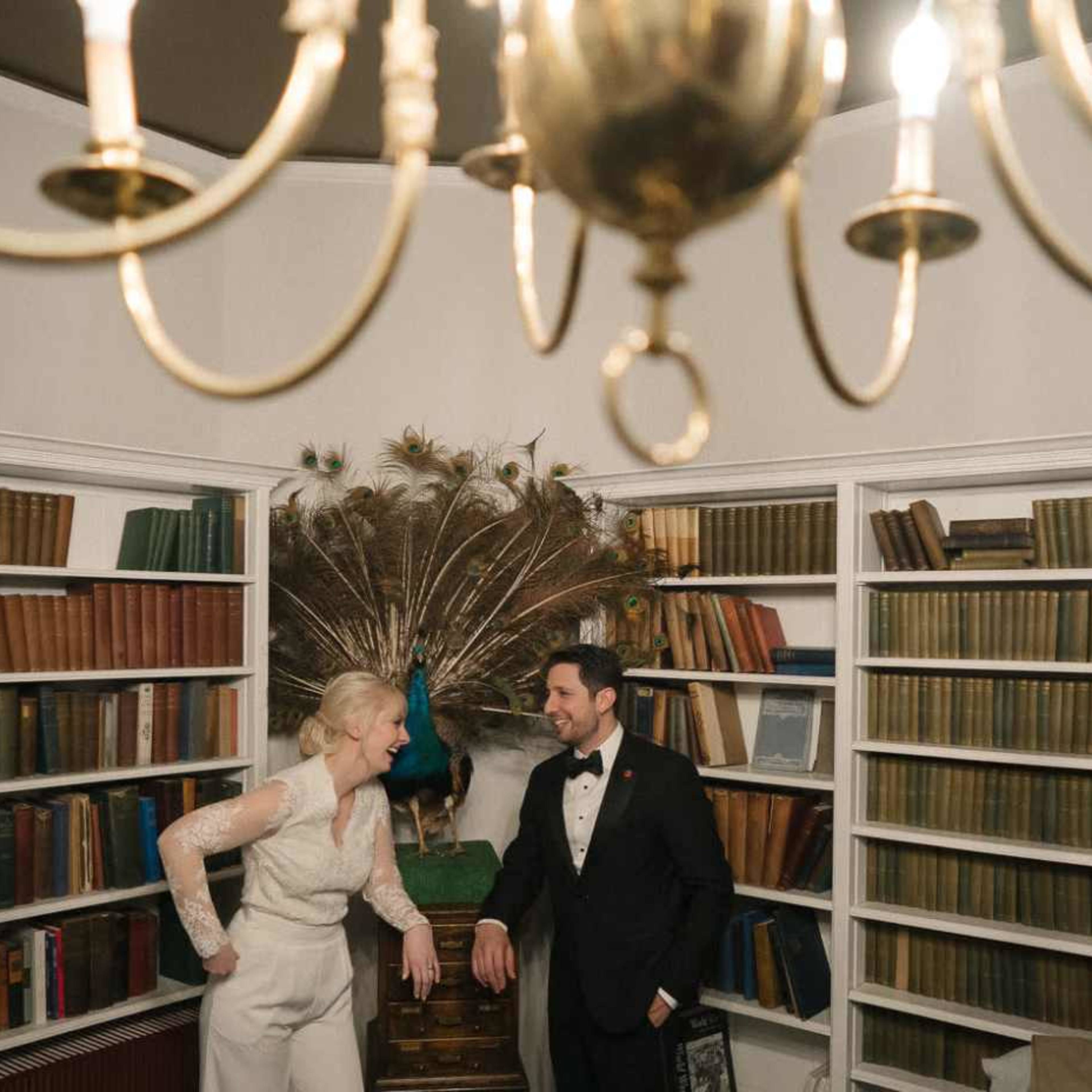 A couple stands in a library with bookshelves filled with various colored books, while a peacock is displayed prominently in the background.