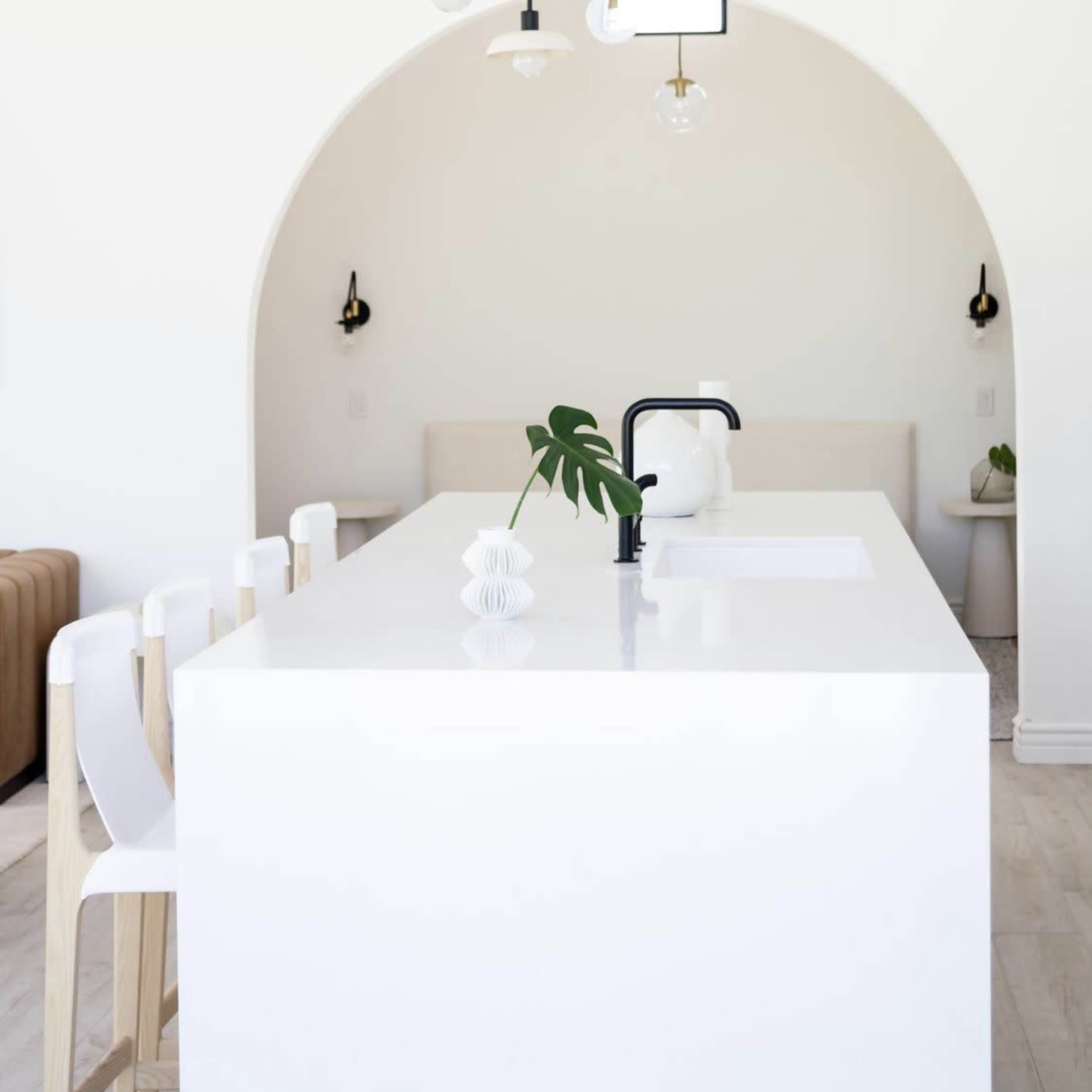 A minimalist kitchen with a large white island, black faucet, and light fixtures, set against an archway leading to a seating area.