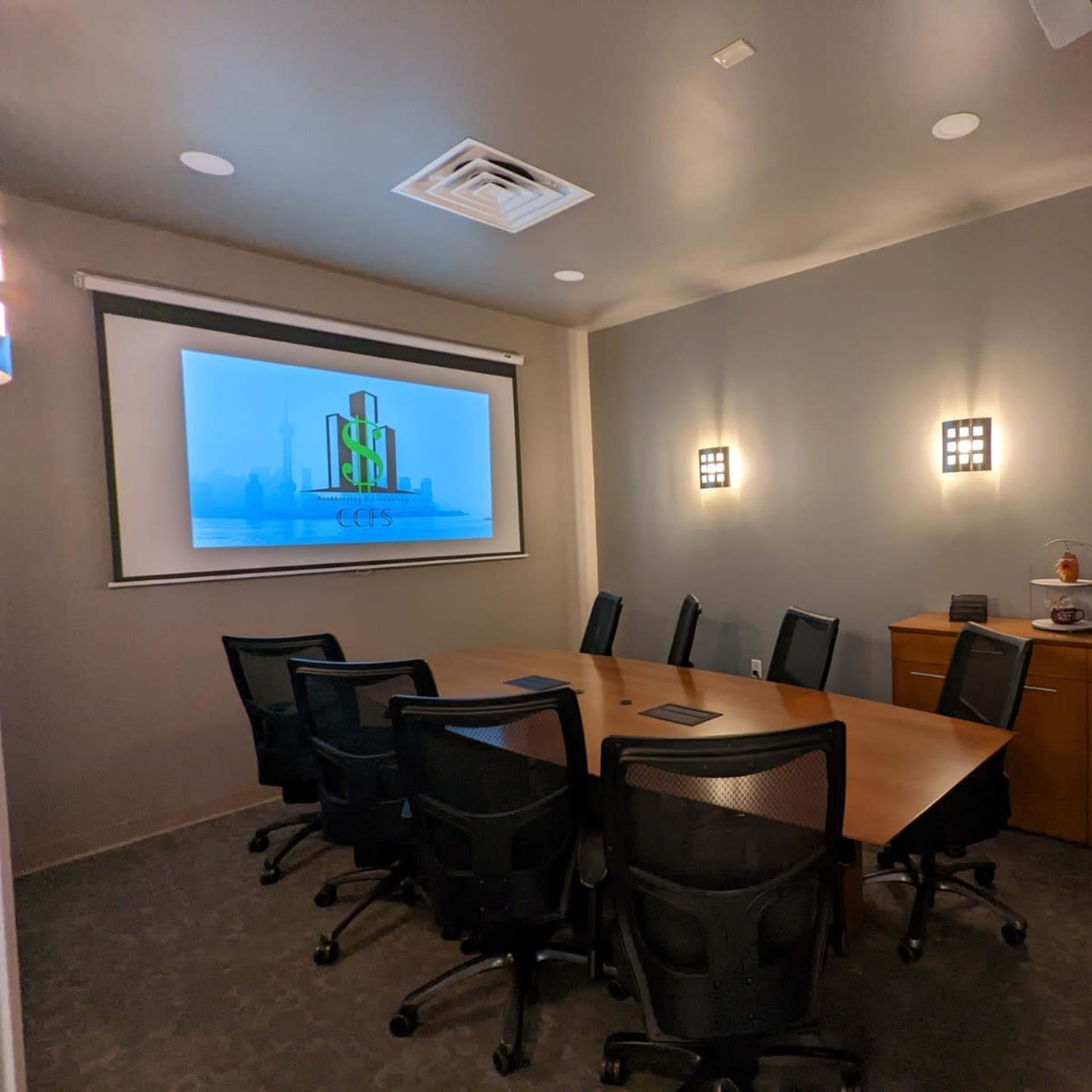 A conference room with a large screen displaying a logo, a wooden table surrounded by black office chairs, and a side cabinet with a coffee machine.