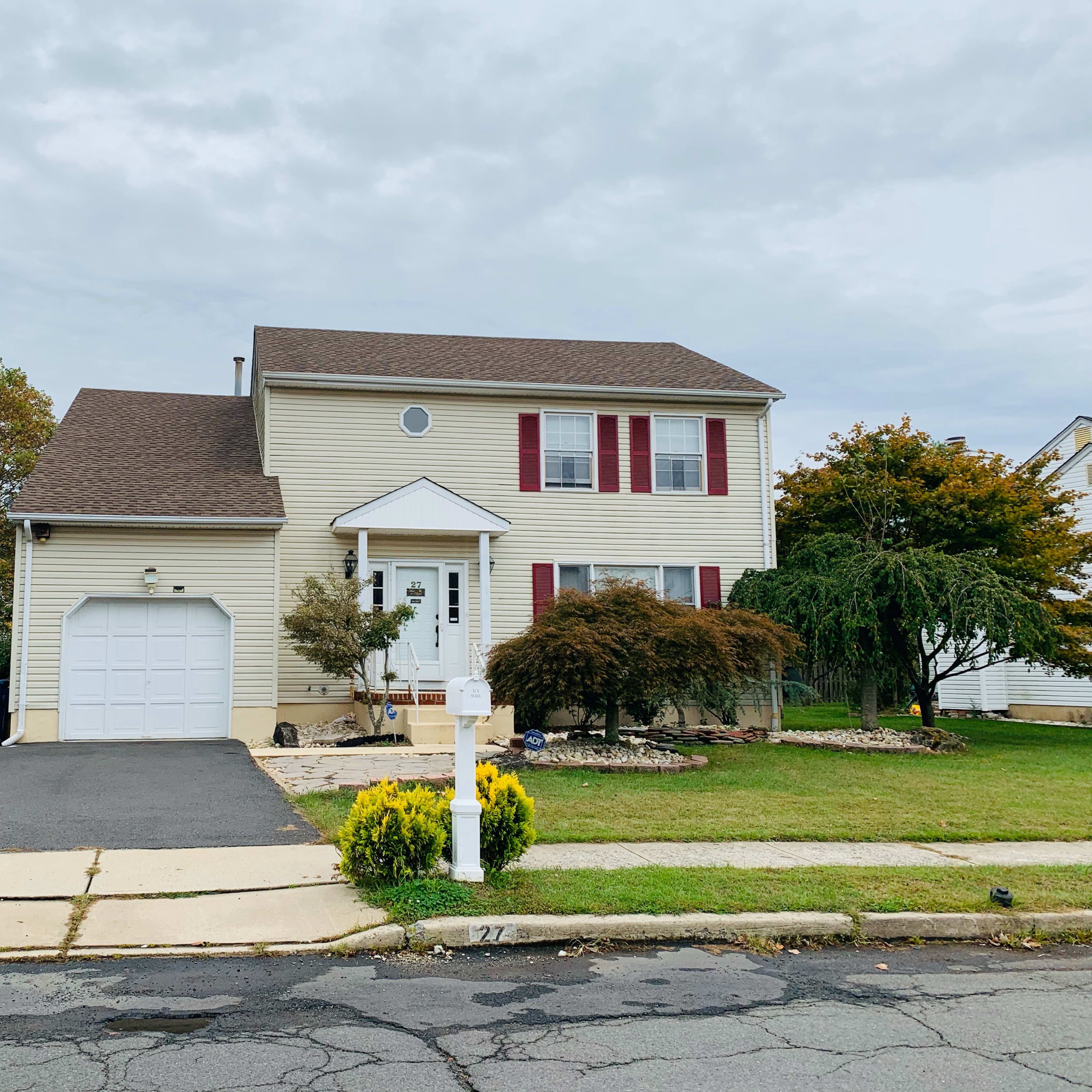 A two-story house with beige siding, red shutters, and a garage is situated on a neatly maintained lawn.