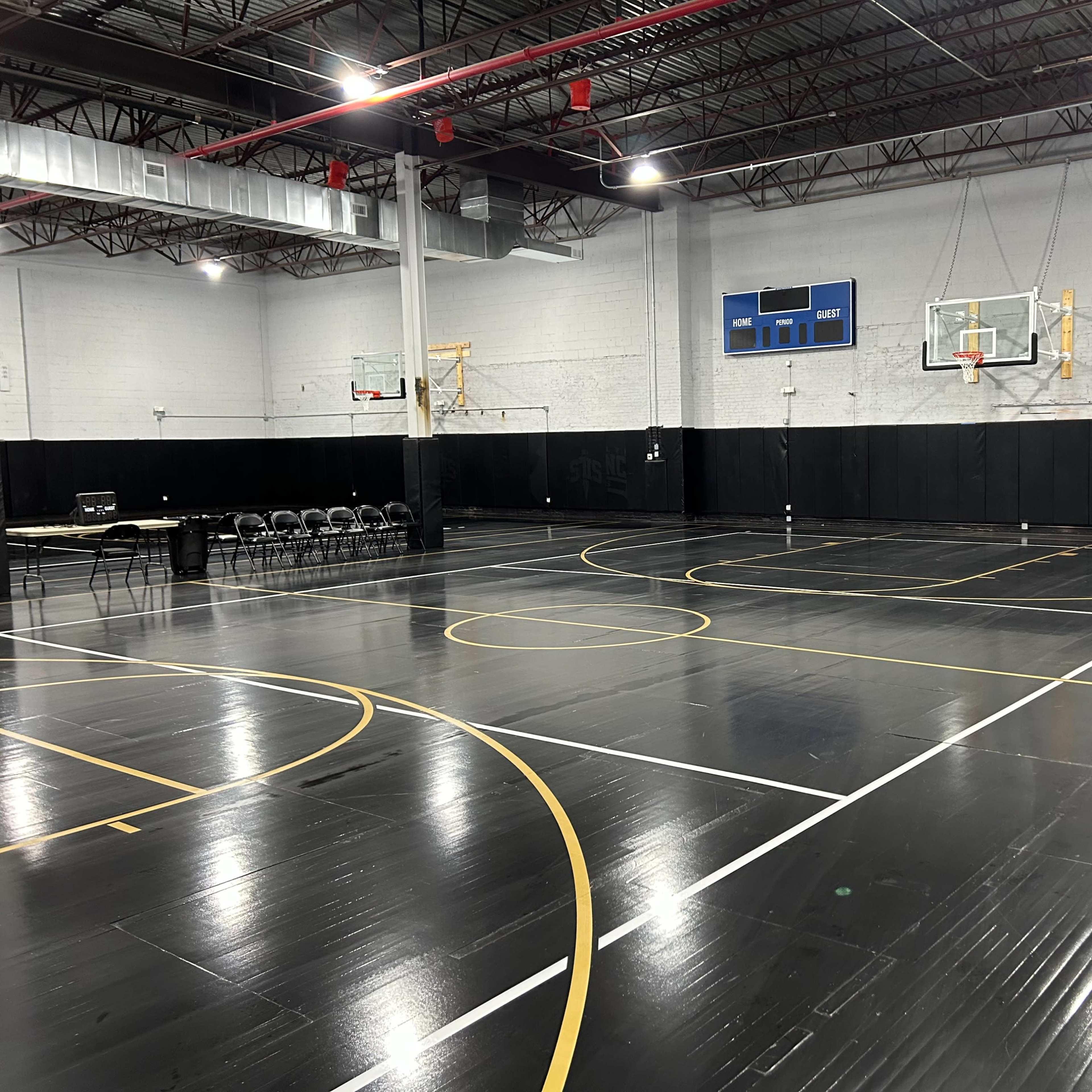 A well-lit indoor basketball court with two hoops, freshly painted lines, and a row of chairs along one side.