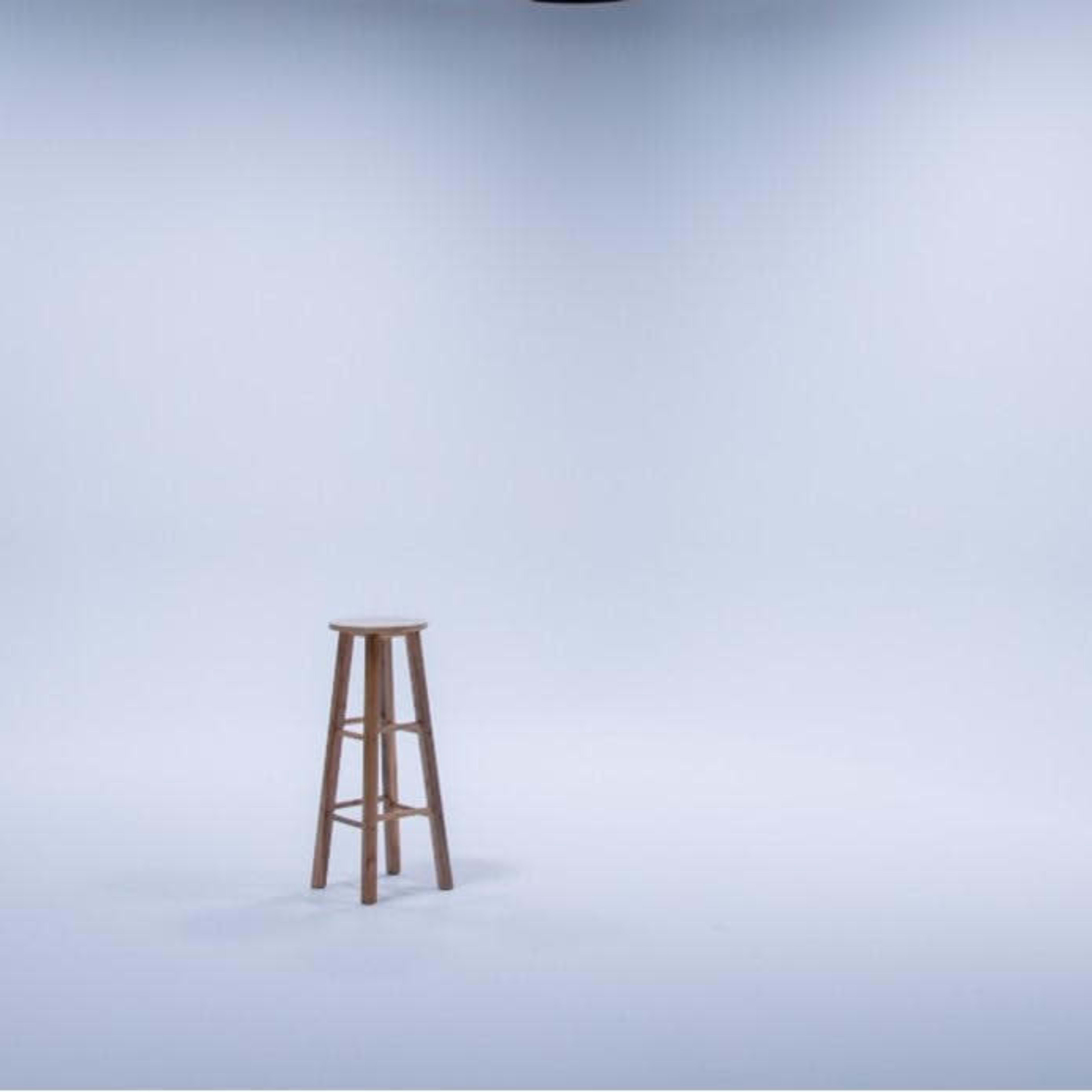 A wooden stool stands alone in a well-lit, empty studio with a light gray background.