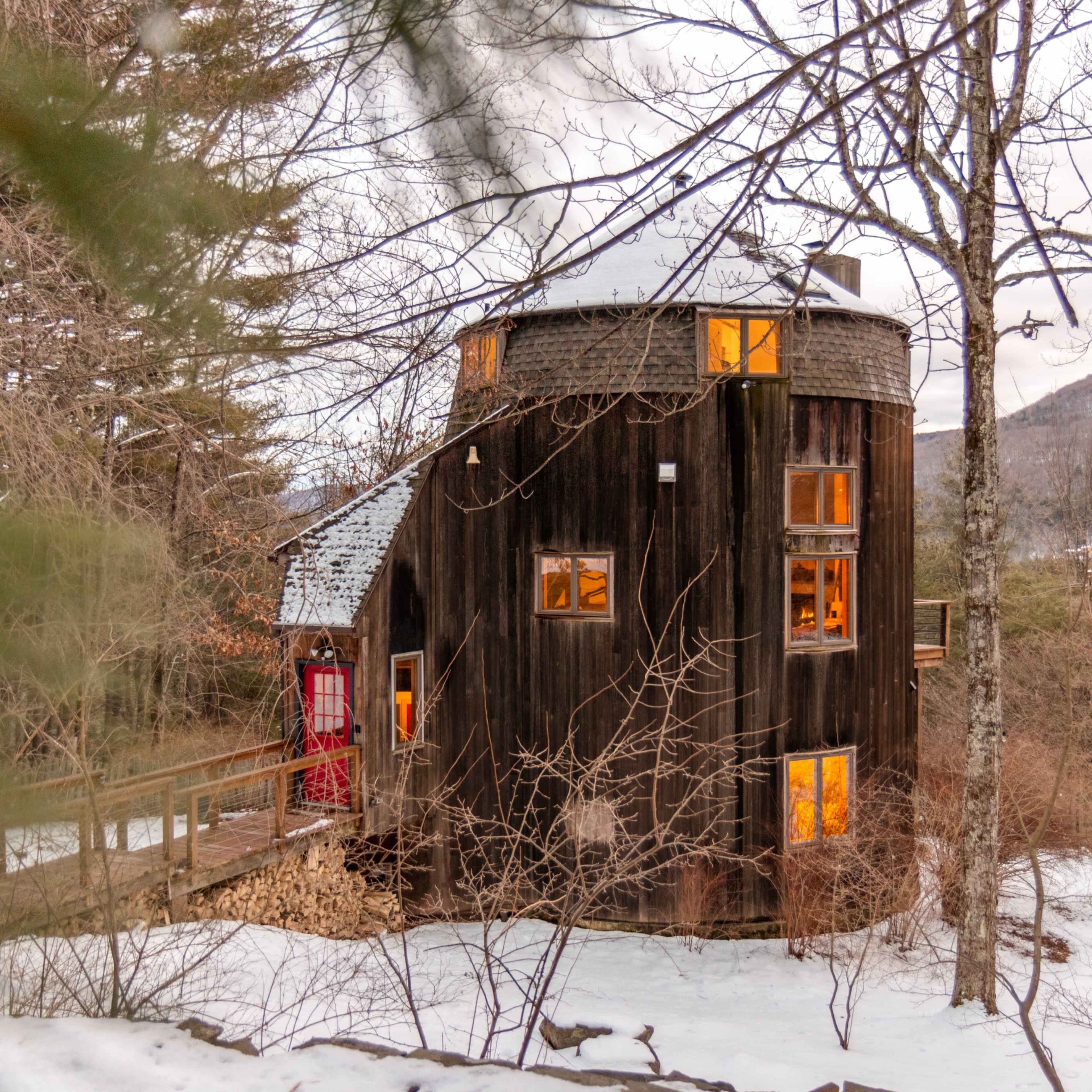 A round, wooden house with large windows is surrounded by trees and snow, set against a backdrop of hills.