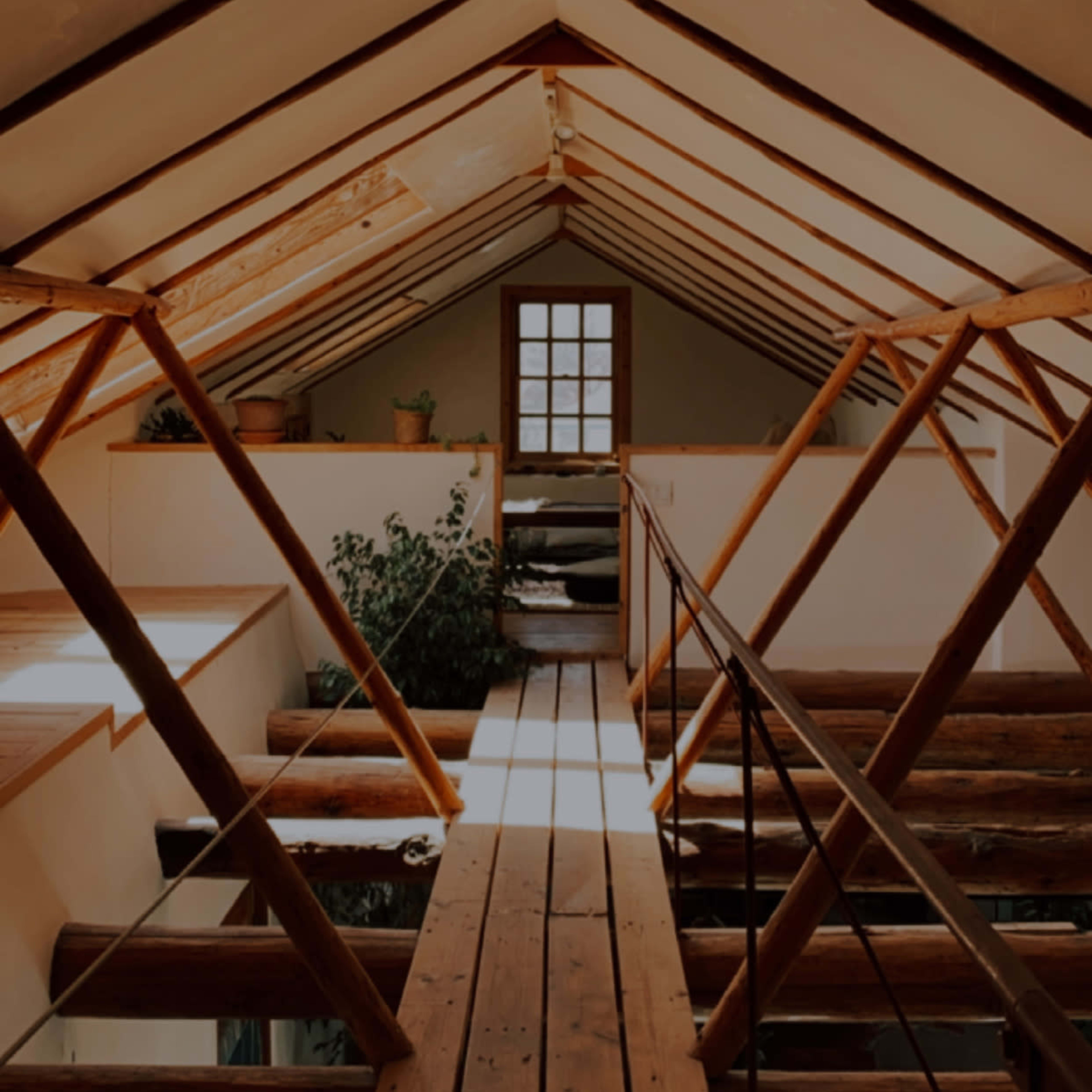 The interior of an attic with exposed wooden beams, a wooden walkway, and greenery near a small window.