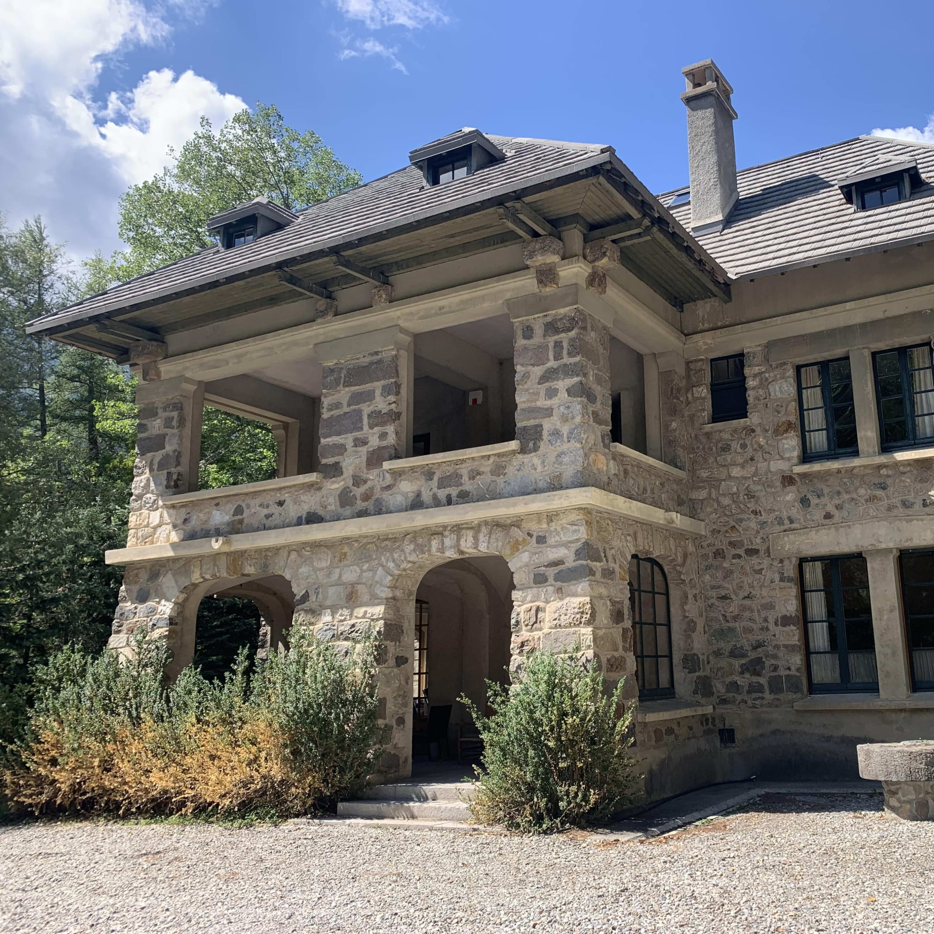 A two-story stone house with a slate roof and large, arched openings on the ground level surrounded by greenery.