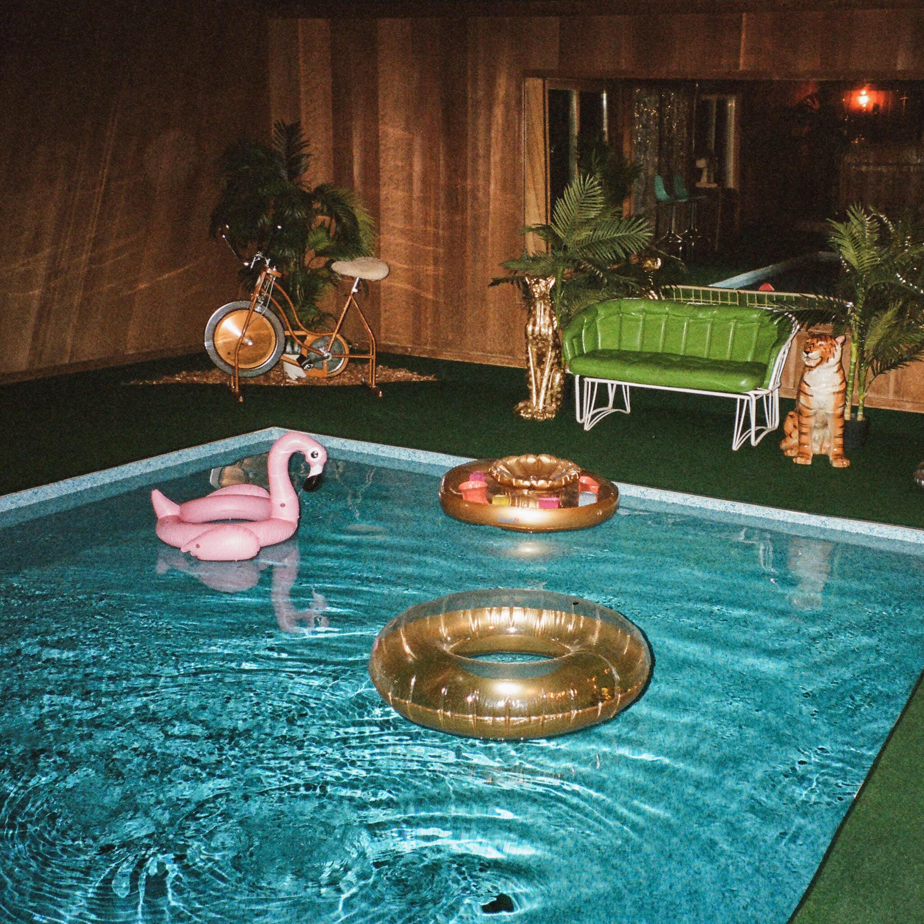 A cozy indoor swimming pool area featuring inflatable pool floats, greenery, and retro furniture.