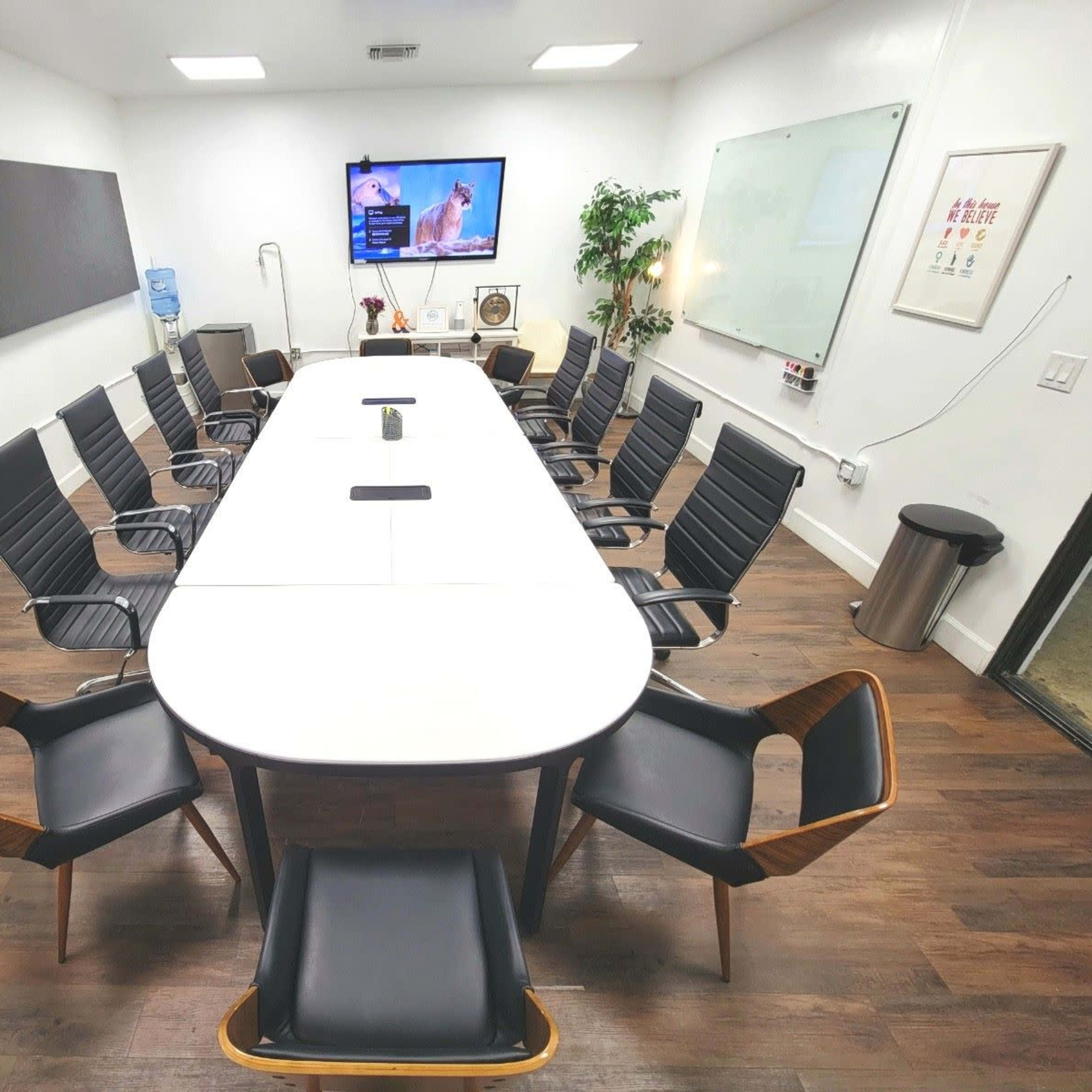 A modern conference room features a large oval table surrounded by black and wood chairs, equipped with a television and whiteboard.