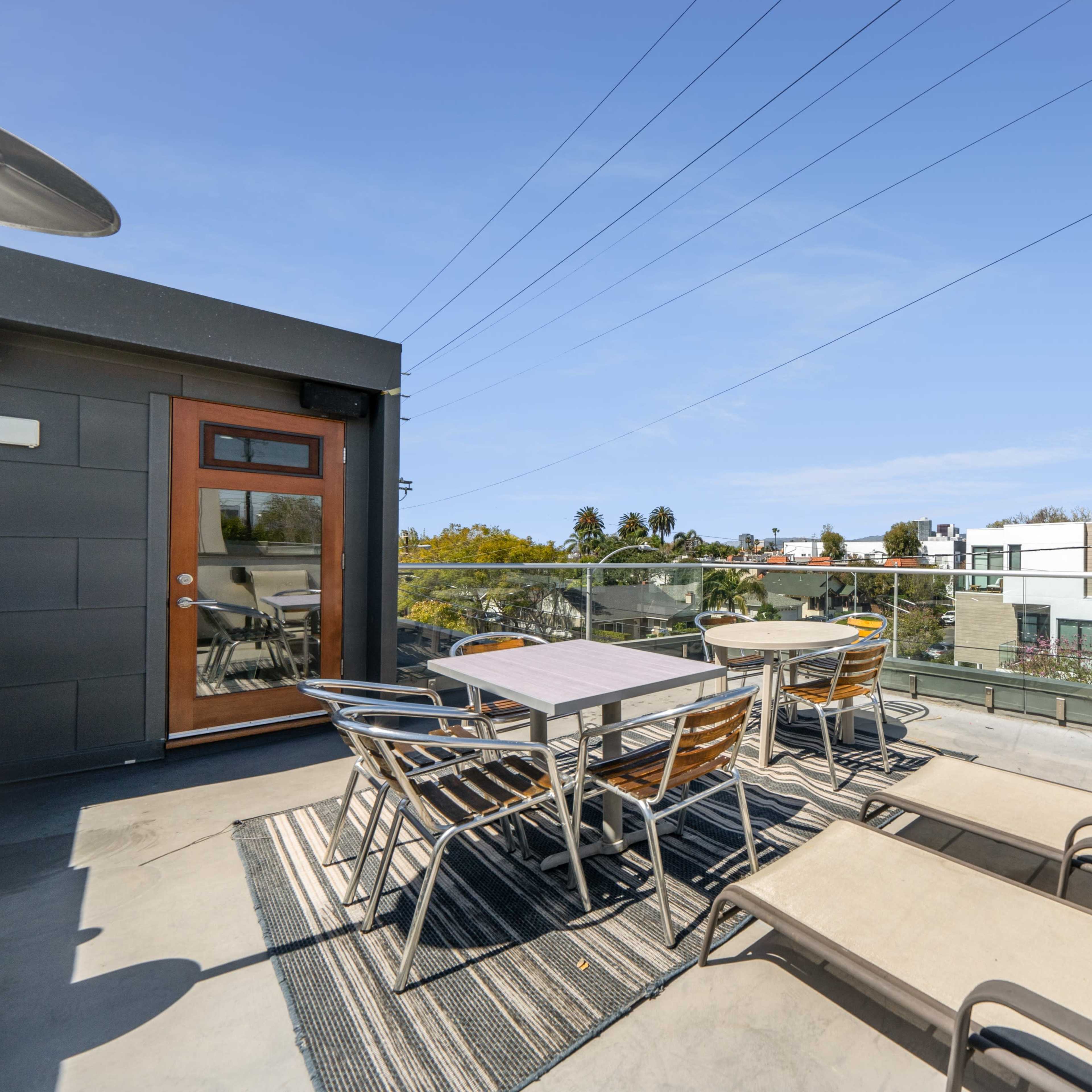 A rooftop patio with tables and lounge chairs, offering a view of nearby buildings and palm trees against a clear sky.