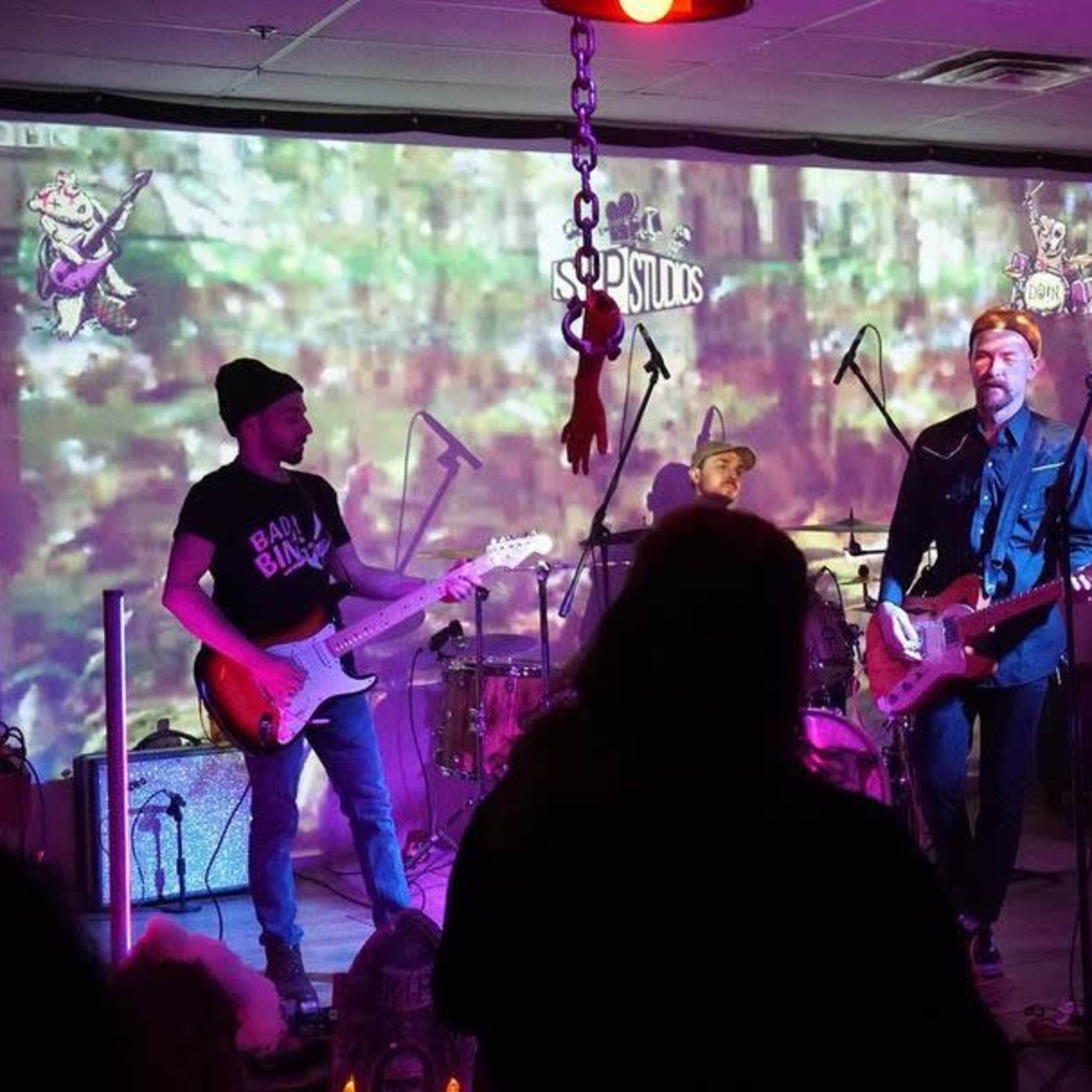 A band performs on stage in a dimly lit venue with a forest-themed backdrop and an audience in front.